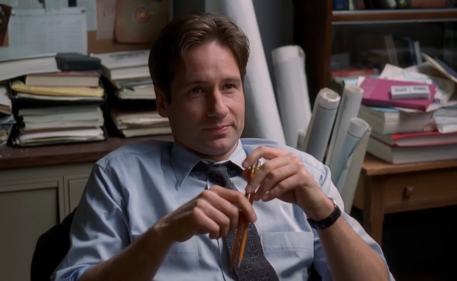 Fox Mulder sitting in an office wearing a blue shirt and tie, holding a pen with architectural plans in the background.