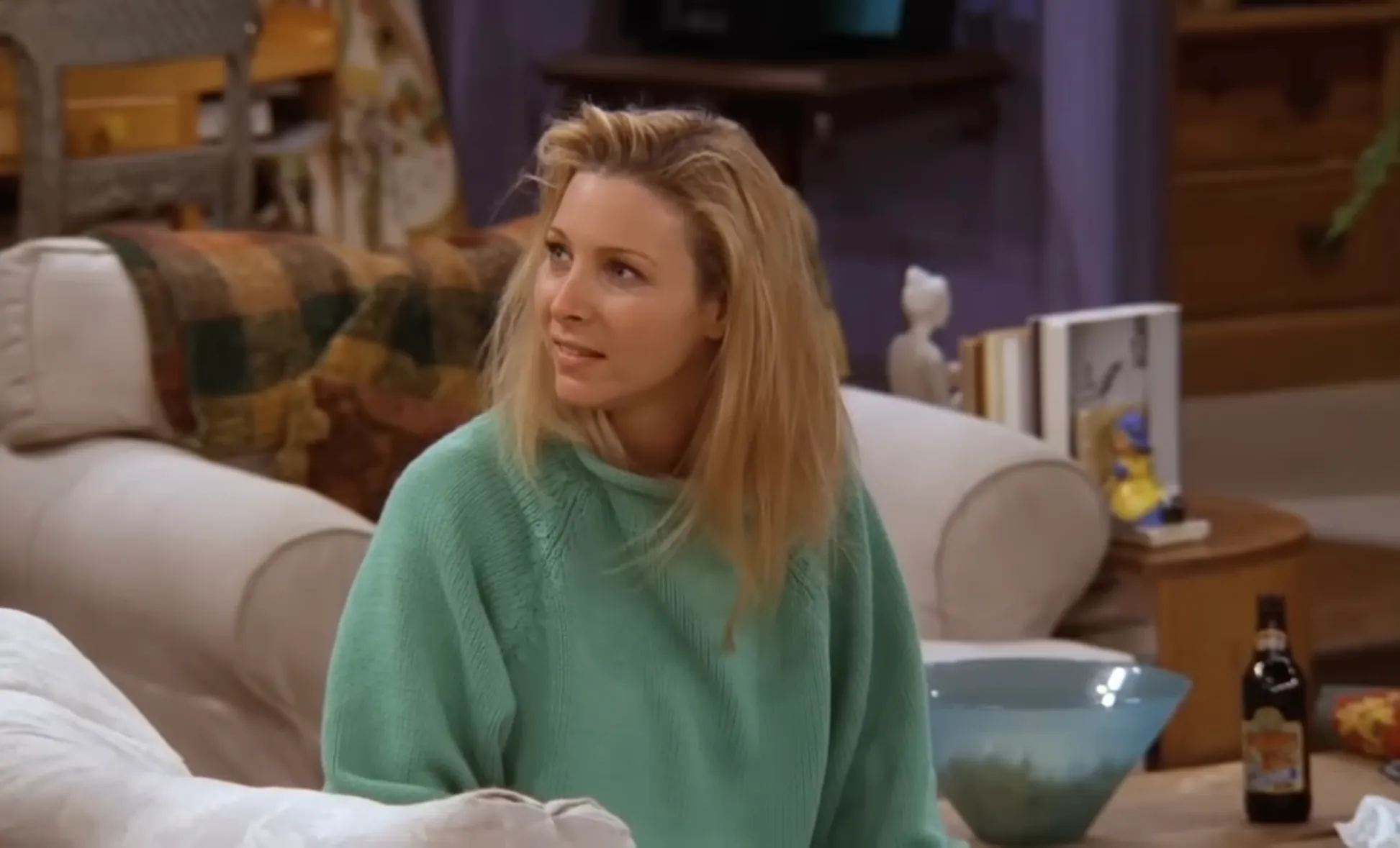 Phoebe from Friends sitting on a couch wearing a green sweater in a living room setting from a TV show.