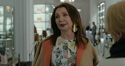 Woman in a floral blouse and beige coat speaking indoors, representing the drama series exploring social media con artist Delvey.
