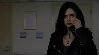 Woman with dark hair and leather jacket holding a phone, looking intense in a dimly lit room, comedy-drama series scene.