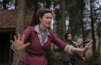A woman in period clothing gestures with hands raised while a man points a gun in the background in a prison drama scene.