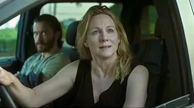 Woman with blonde hair sitting in a car, speaking, with a man in the passenger seat behind her in a prison drama scene.