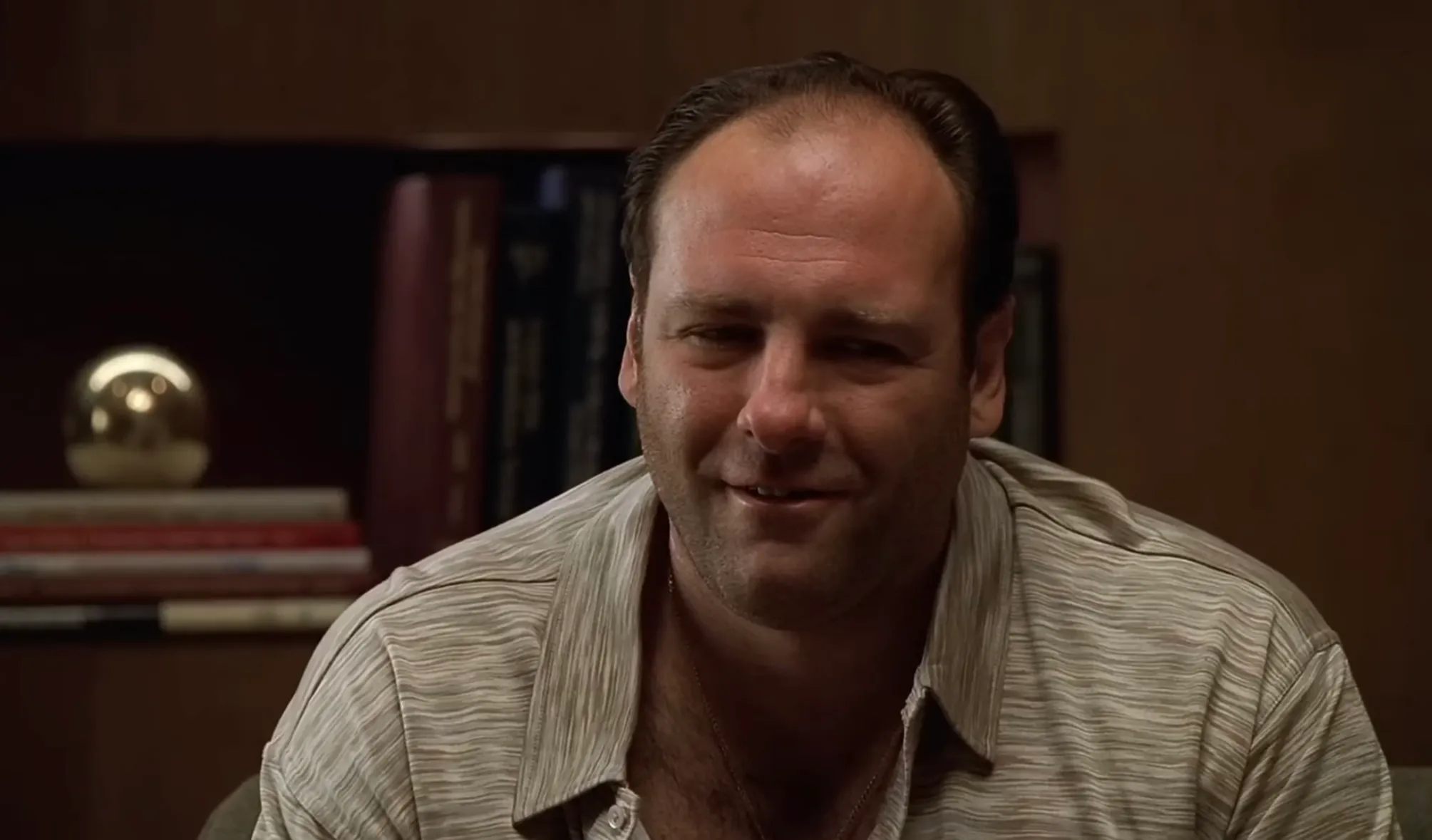 Tony Soprano smiling in a striped shirt, with books and a golden sphere on a shelf in the background.