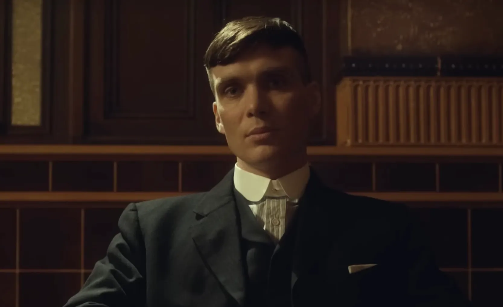 Thomas Shelby sitting in a dark room wearing a black suit and white collar, looking directly at the camera.