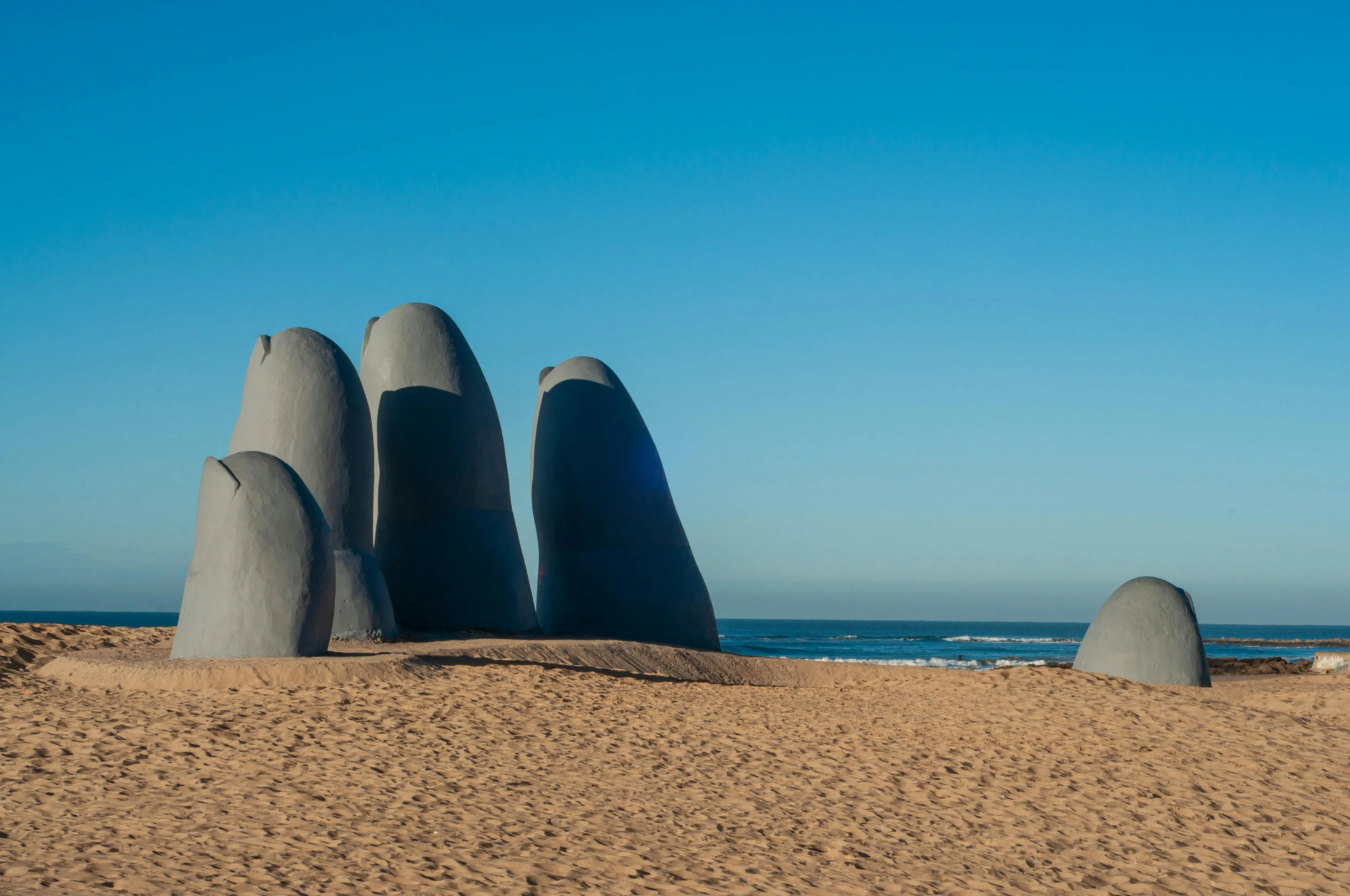 Large stone hand sculpture emerging from sand on a beach under blue sky, part of guess 25 countries single picture challenge.