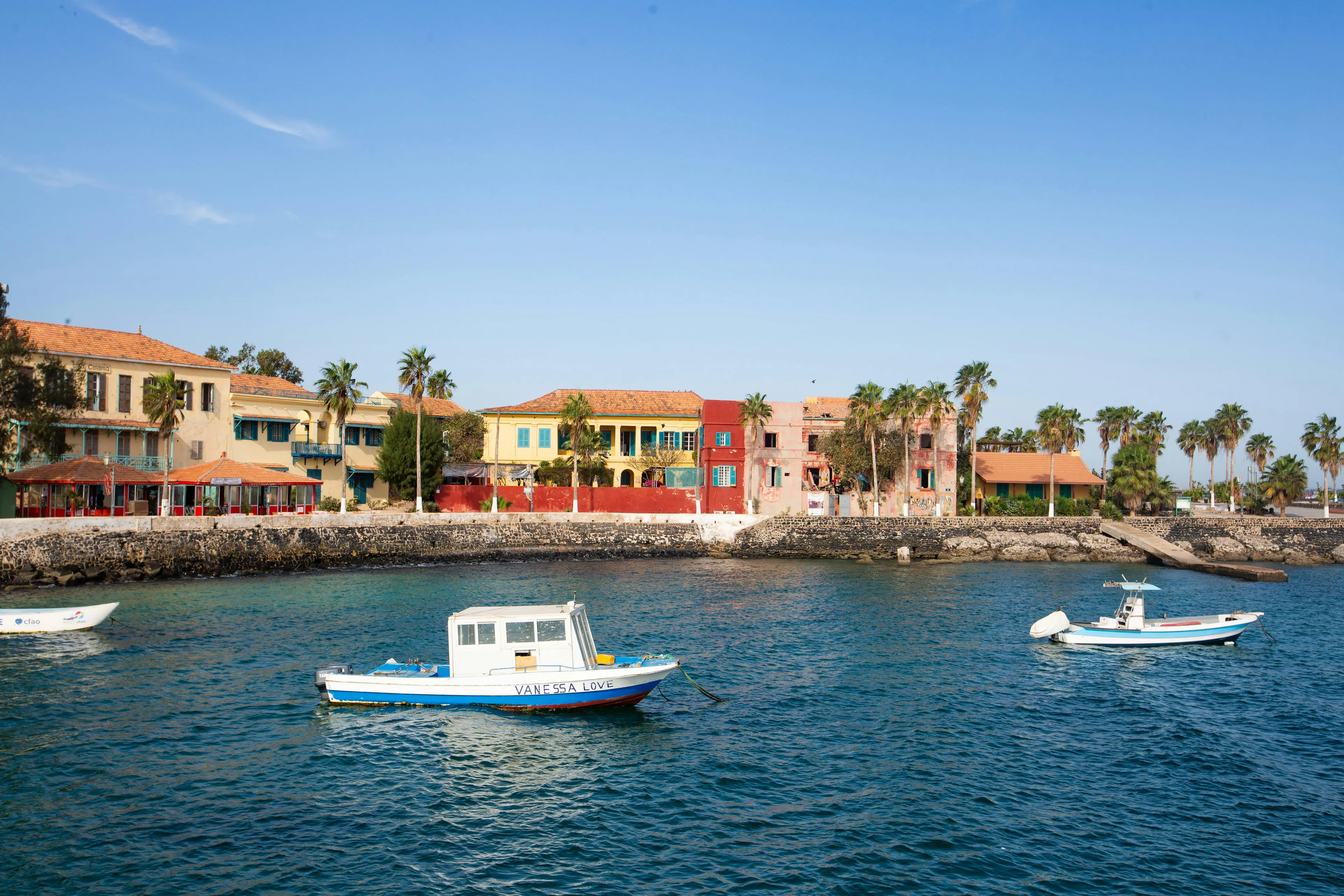 Coastal scene in a West African country with fishing boats, colorful buildings, and palm trees under a clear sky.