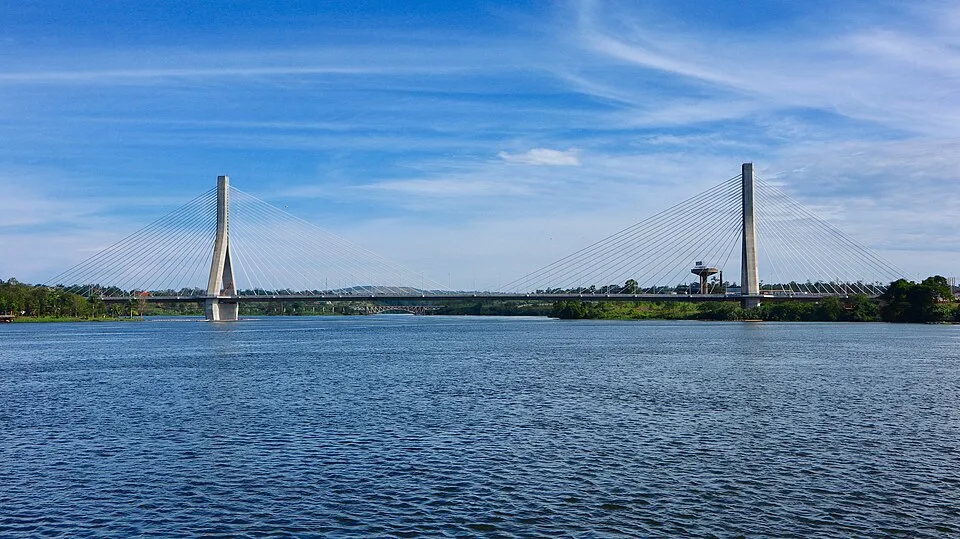 Suspension bridge over calm river under blue sky, inviting viewers to guess the country from a single picture.
