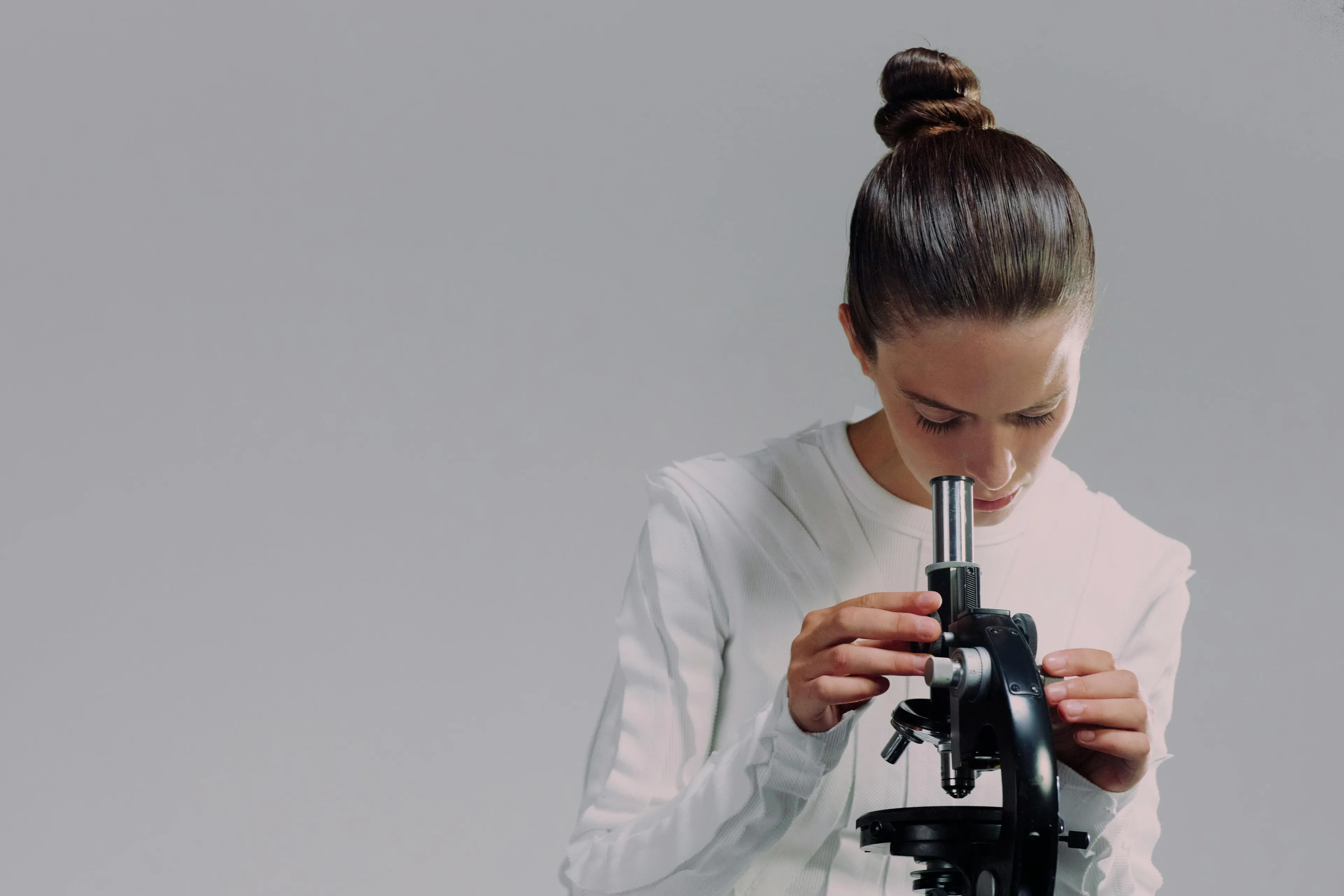 Young woman in white shirt looking through a black microscope, engaging in science and learning about element with atomic number 12.