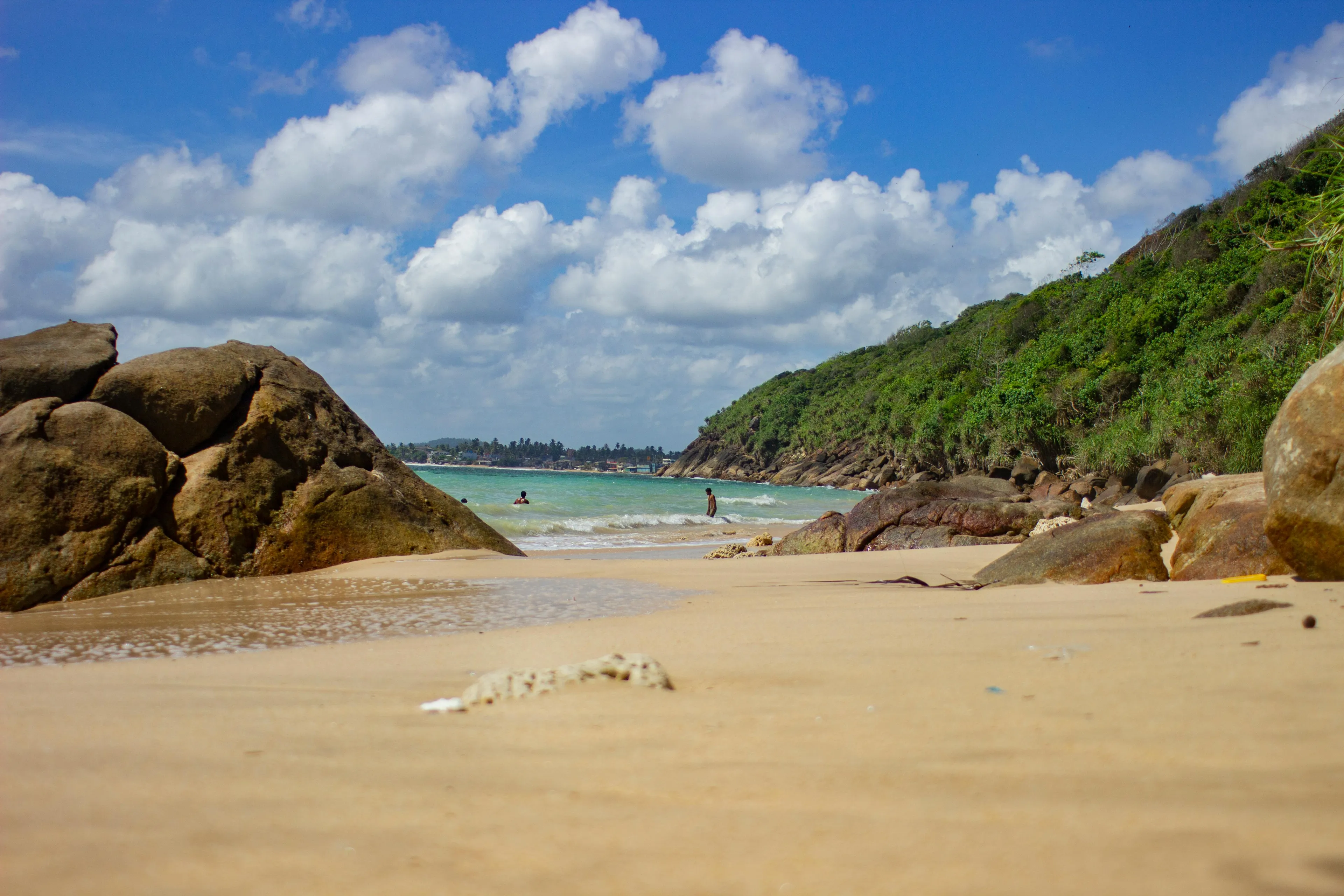 Beach view in Sri Lanka with rocks, sandy shore, green hillside, and people swimming under a partly cloudy sky.