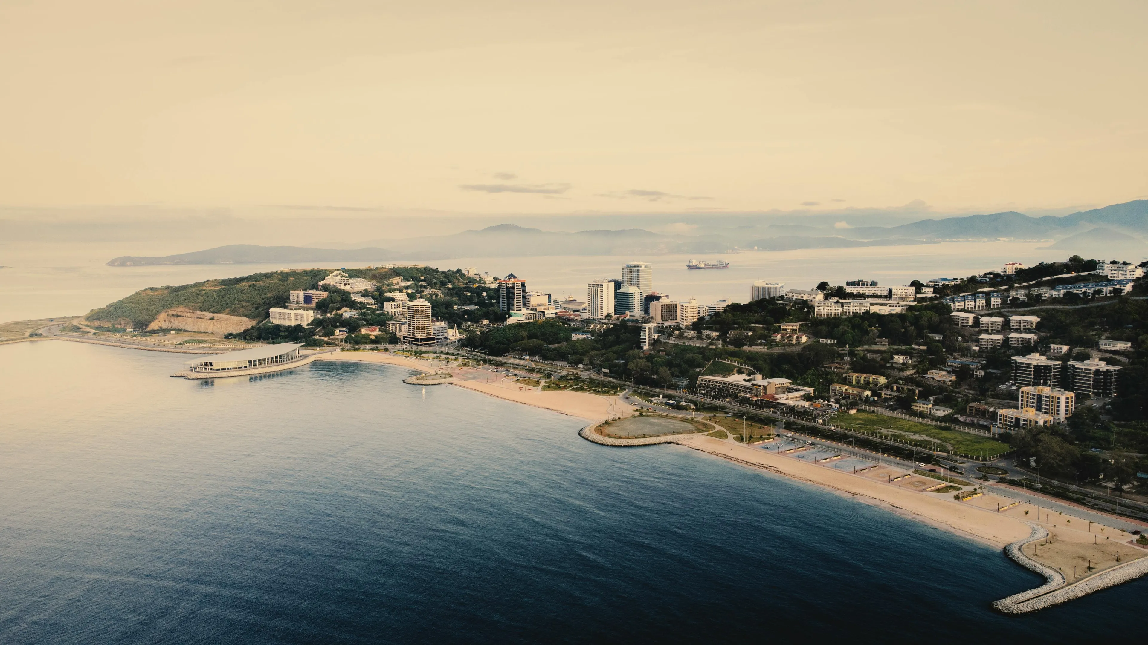 Coastal cityscape of Papua New Guinea with beachfront, urban buildings, and mountains along the Pacific Ocean shore.