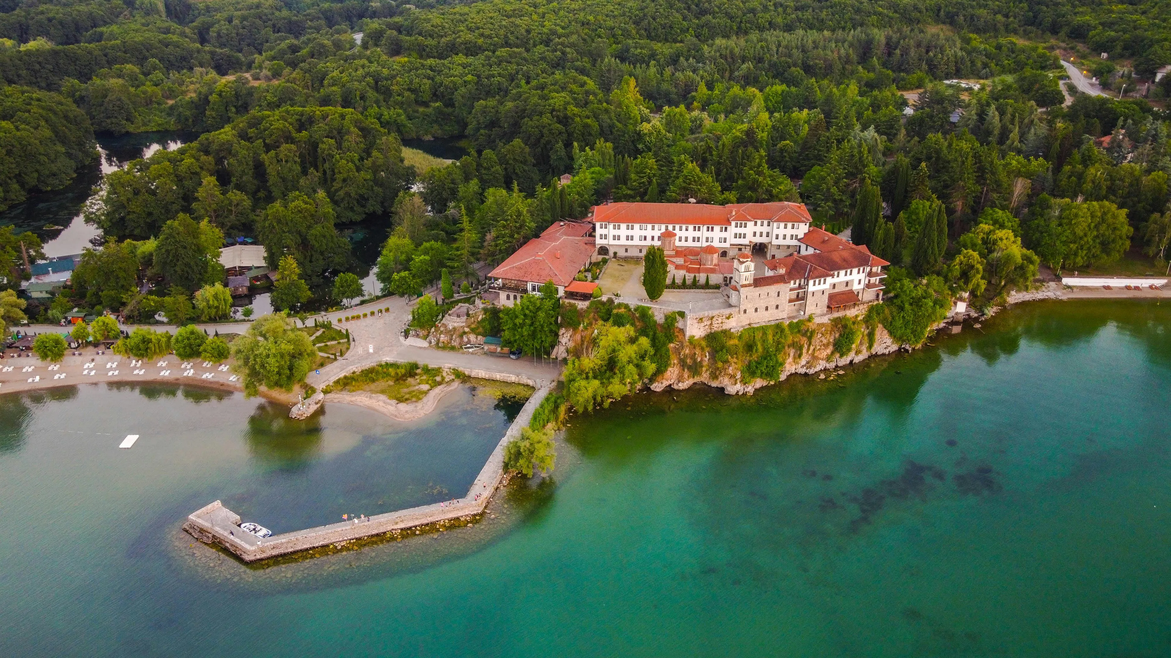 Aerial view of a historic monastery on a cliff surrounded by forest and turquoise water in North Macedonia.