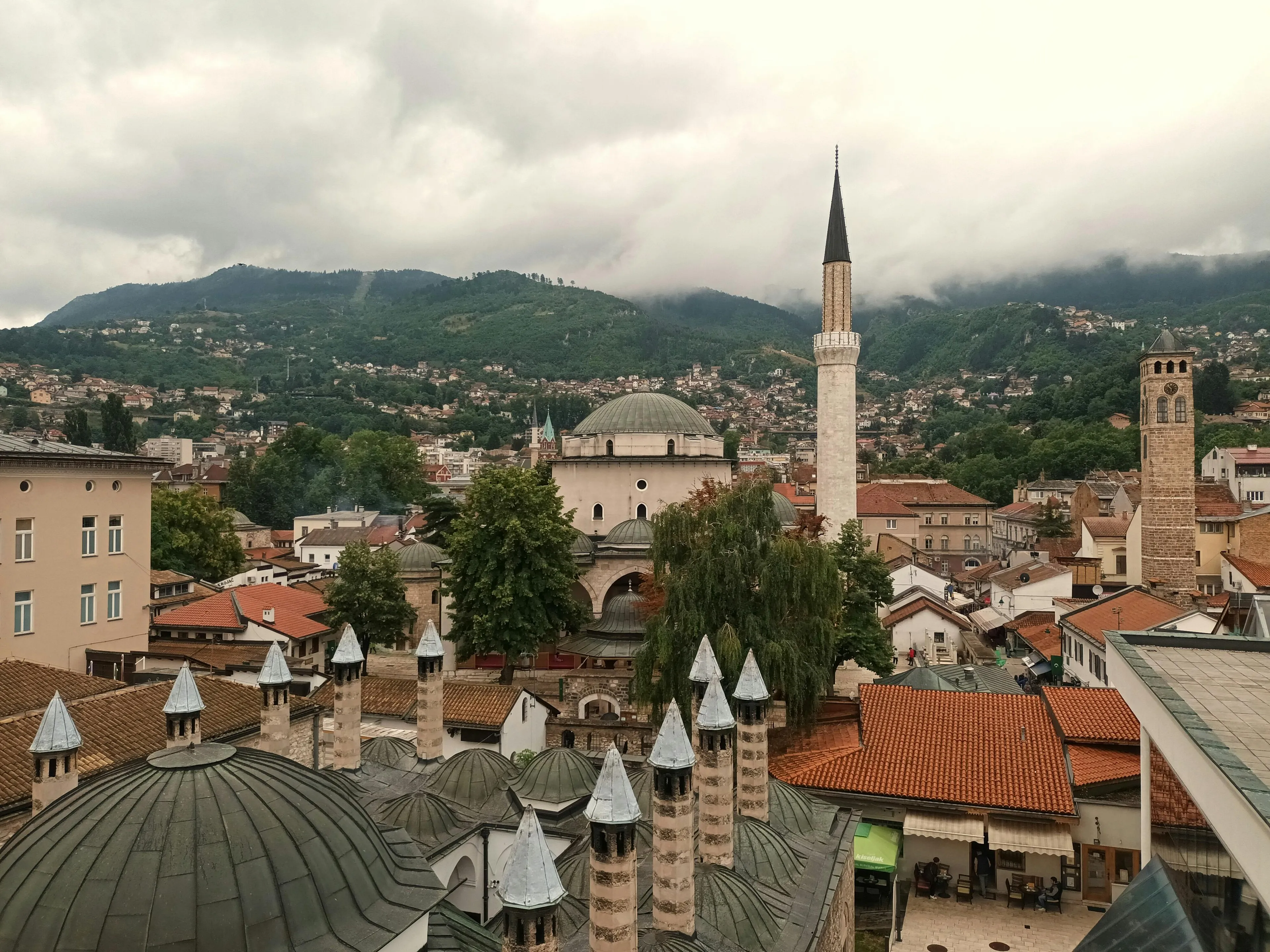 View of historic buildings and mosques in Bosnia and Herzegovina with mountainous background under cloudy sky.