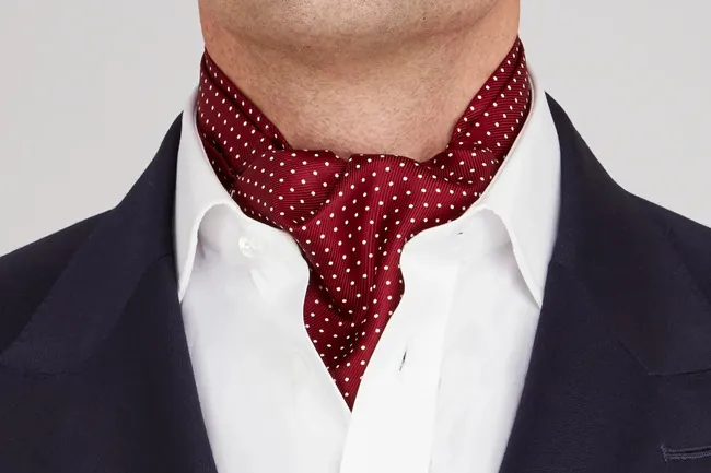 Close-up of a man wearing a silk ascot with white polka dots, paired with a white shirt and dark blazer.