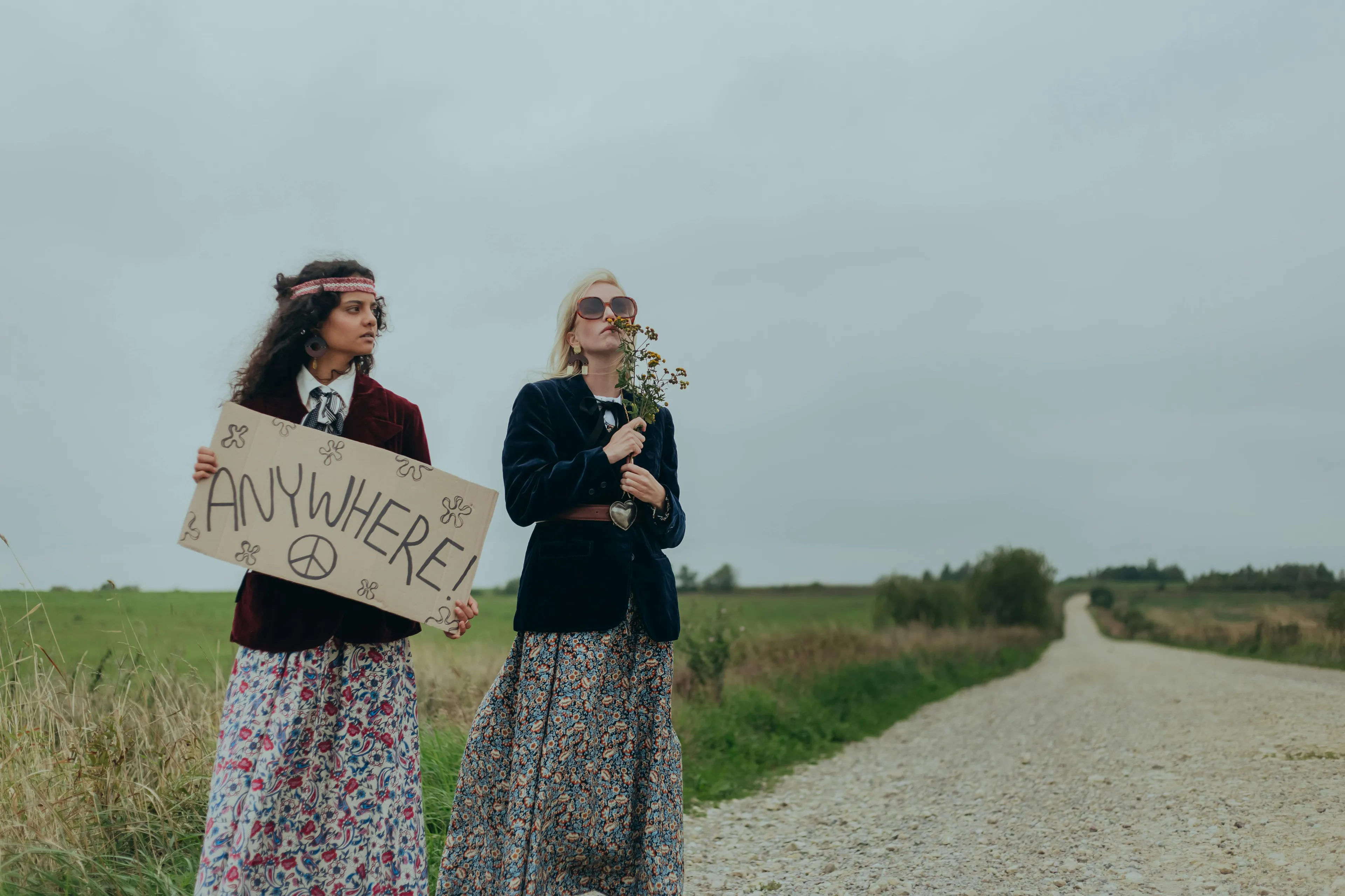 Two women in floral skirts stand on a rural road holding sign with anywhere text, representing dream vacation spot choices.