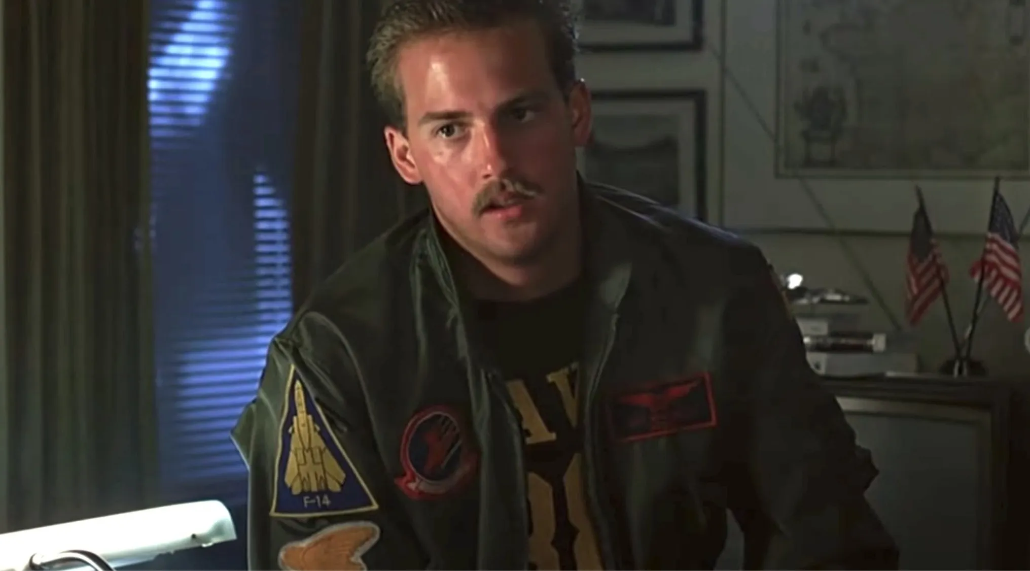 Side character from Top Gun wearing a military jacket with patches sitting in a dimly lit room with American flags.