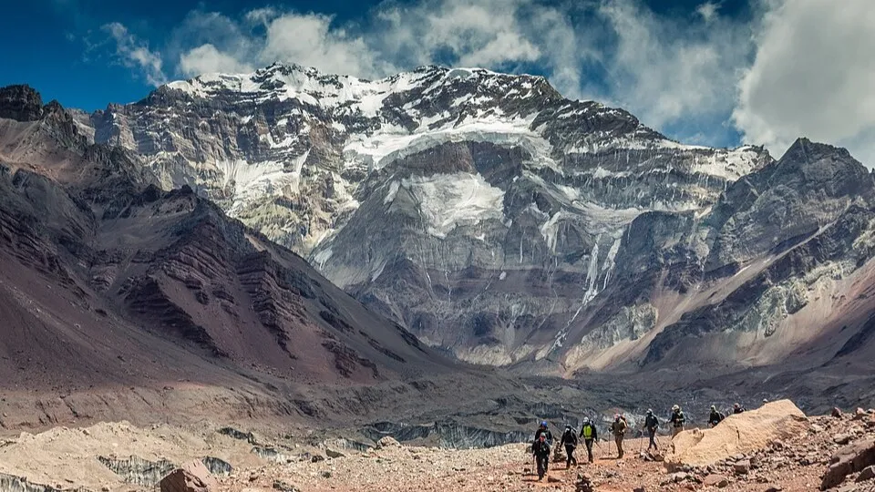 Hikers approaching the rocky and snowy summit of Mount Aconcagua in Argentina under a partly cloudy sky.