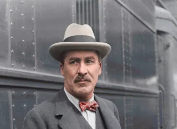 British archaeologist Howard Carter wearing a hat and bow tie standing next to a metal train carriage.