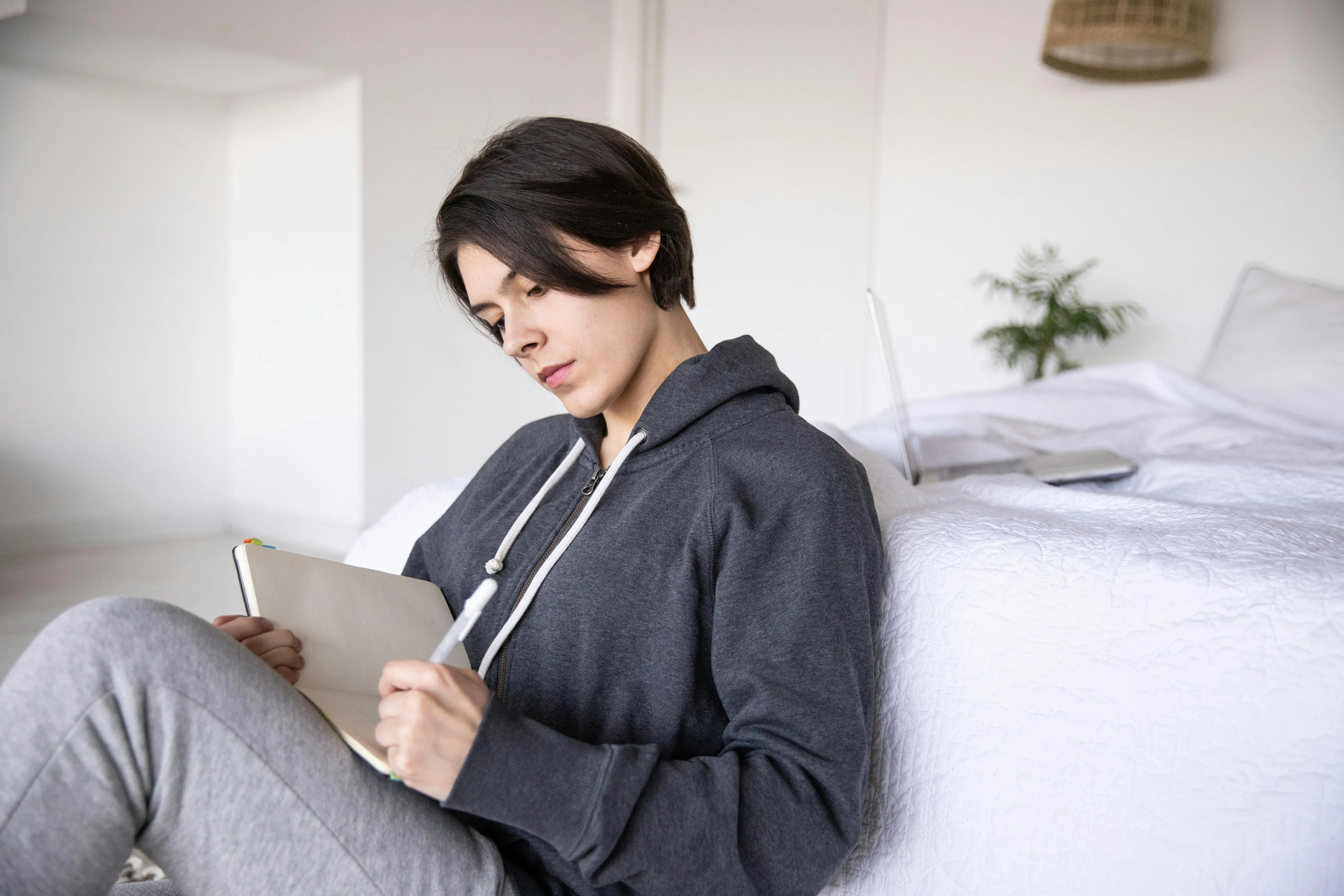 Person in casual clothes sitting by bed and writing on a tablet, reflecting on after-work ritual habits for sitcom quiz.