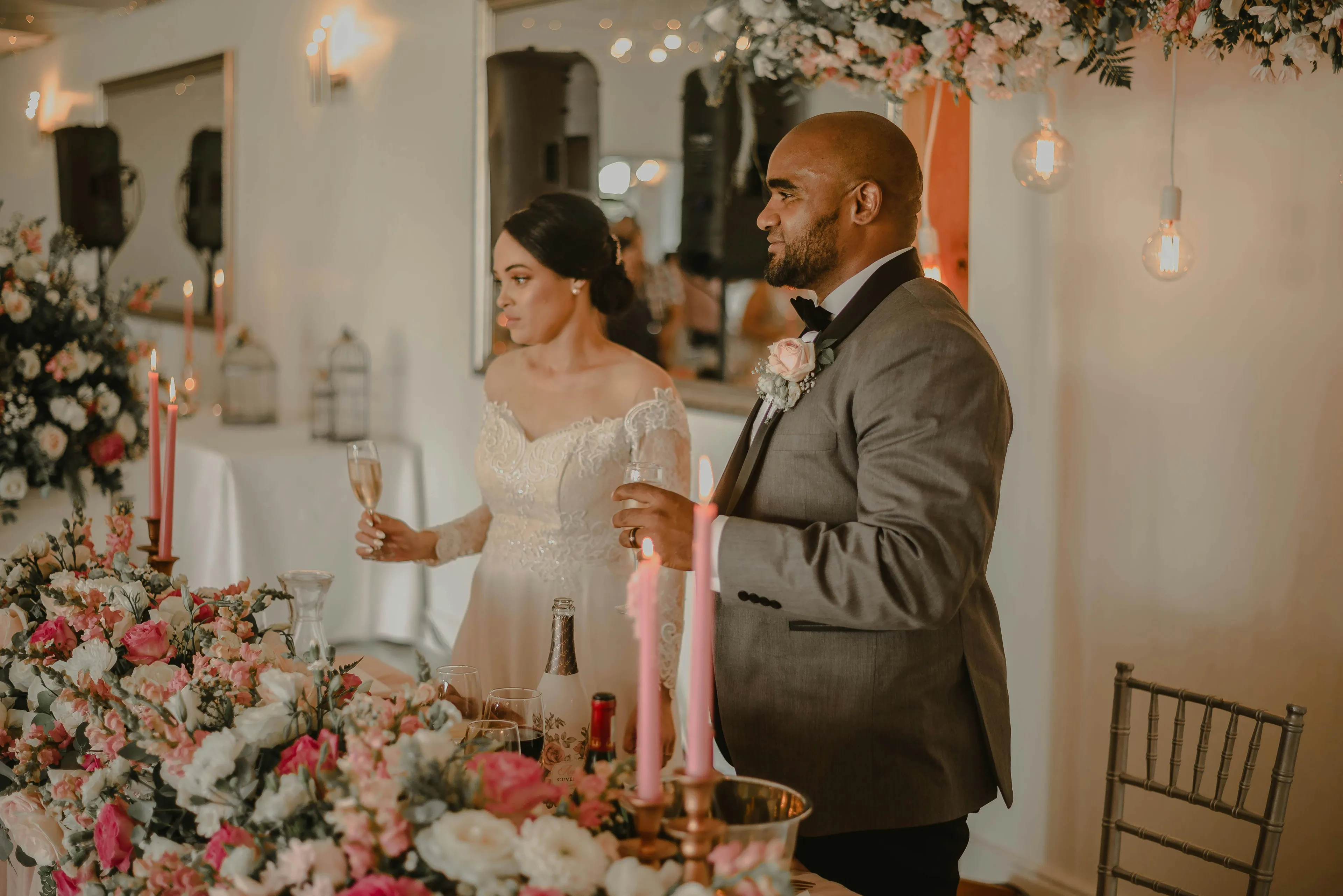 Couple raising glasses at a wedding reception surrounded by floral decorations and warm lighting, sitcom setting ambiance.