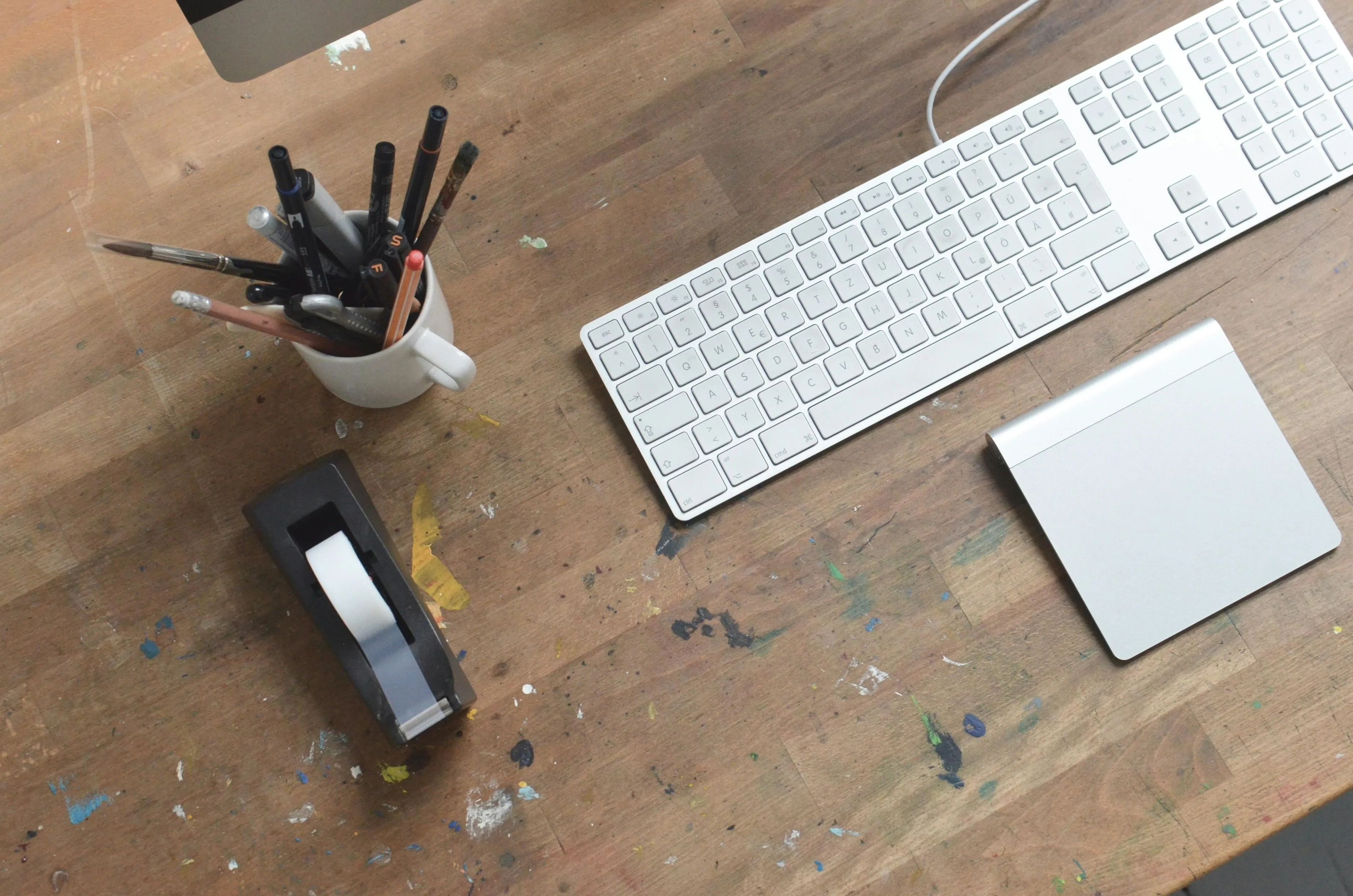 Workspace organization with pens in a cup, tape dispenser, keyboard, and trackpad on a wooden desk surface.