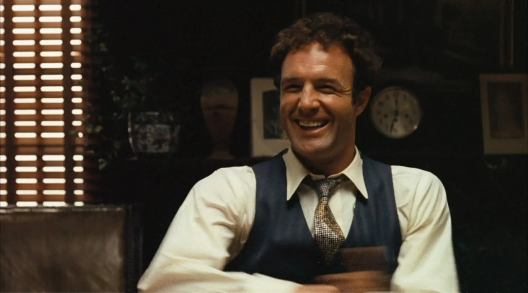 Side character from a movie, hot-tempered and impulsive, smiling in a vintage setting with a clock and window blinds.