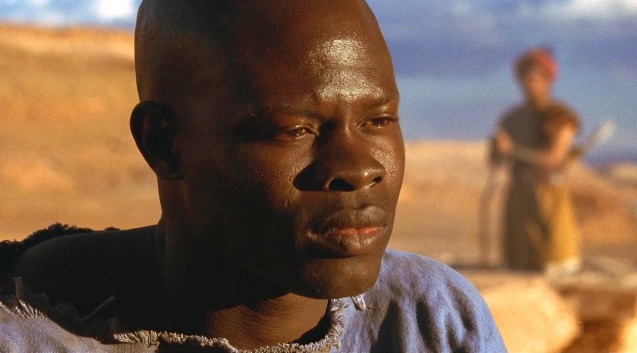 Side character from Gladiator with close-up on face wearing a blue garment in a desert setting with blurred background.