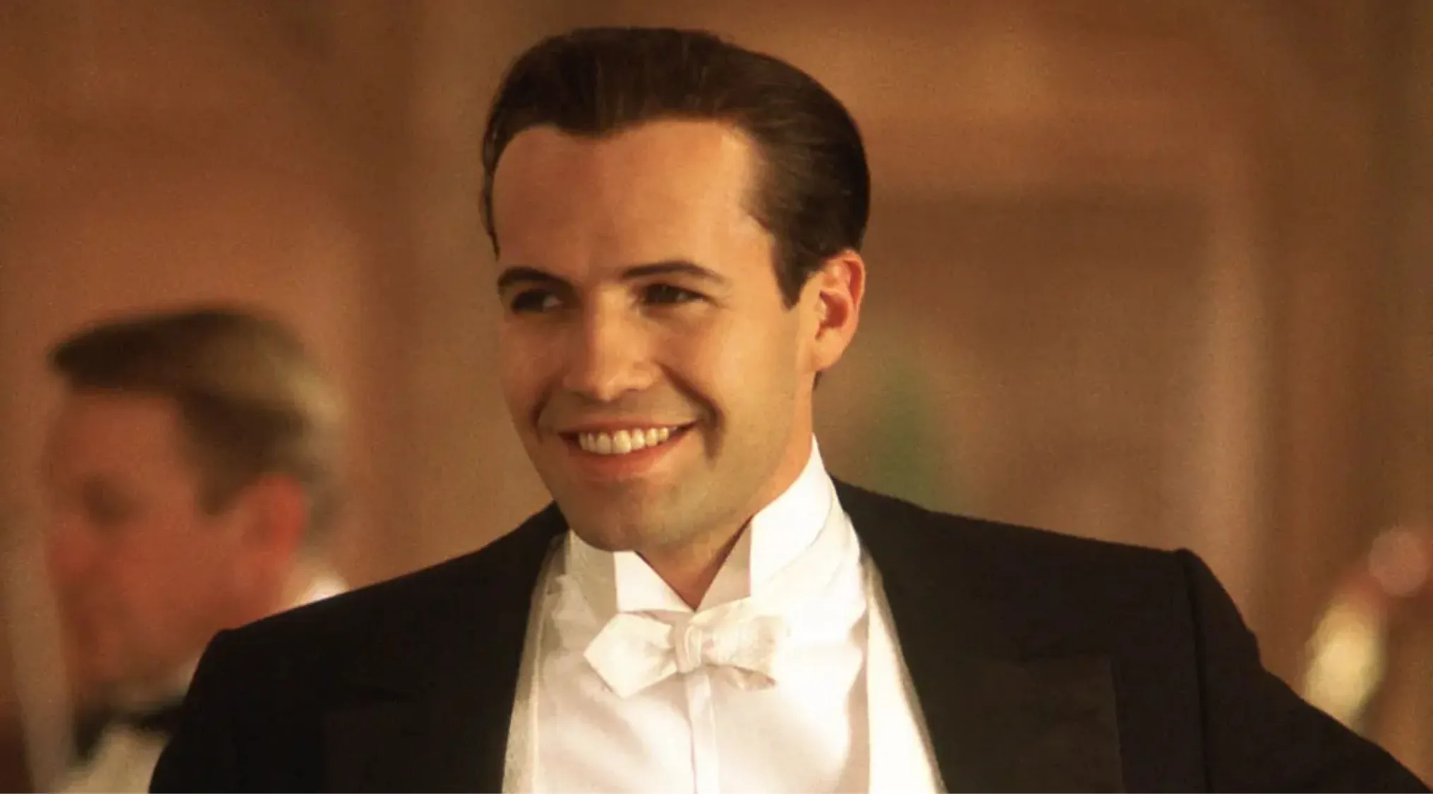 Billy Zane smiling in a black tuxedo and white bow tie, portraying a side character in Titanic movie scene.