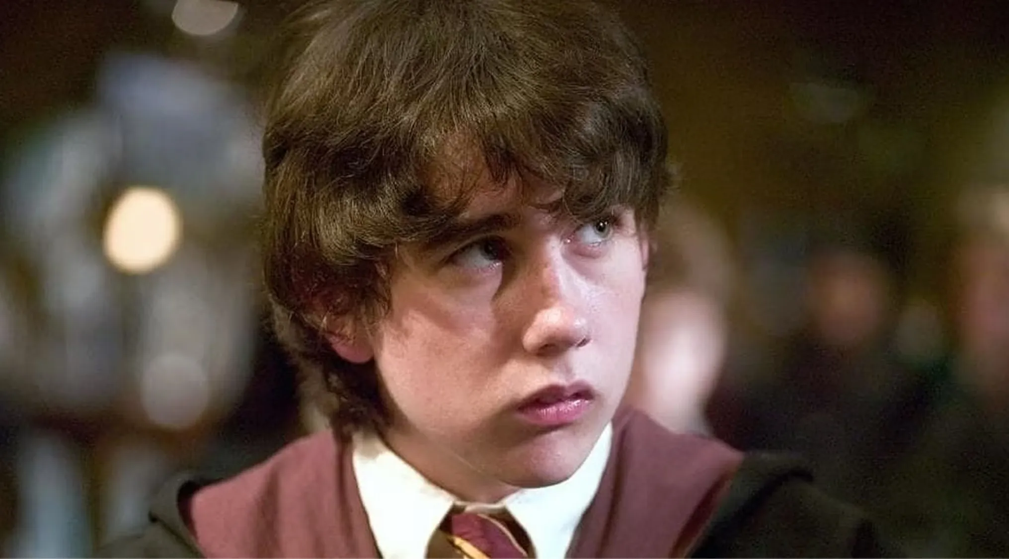 Young side character from Harry Potter movies wearing a Gryffindor uniform looking to the side in a dimly lit setting