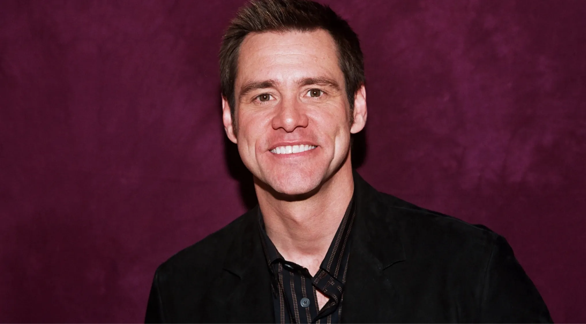 Jim Carrey smiling in a black jacket against a dark purple background in a movie side character role.