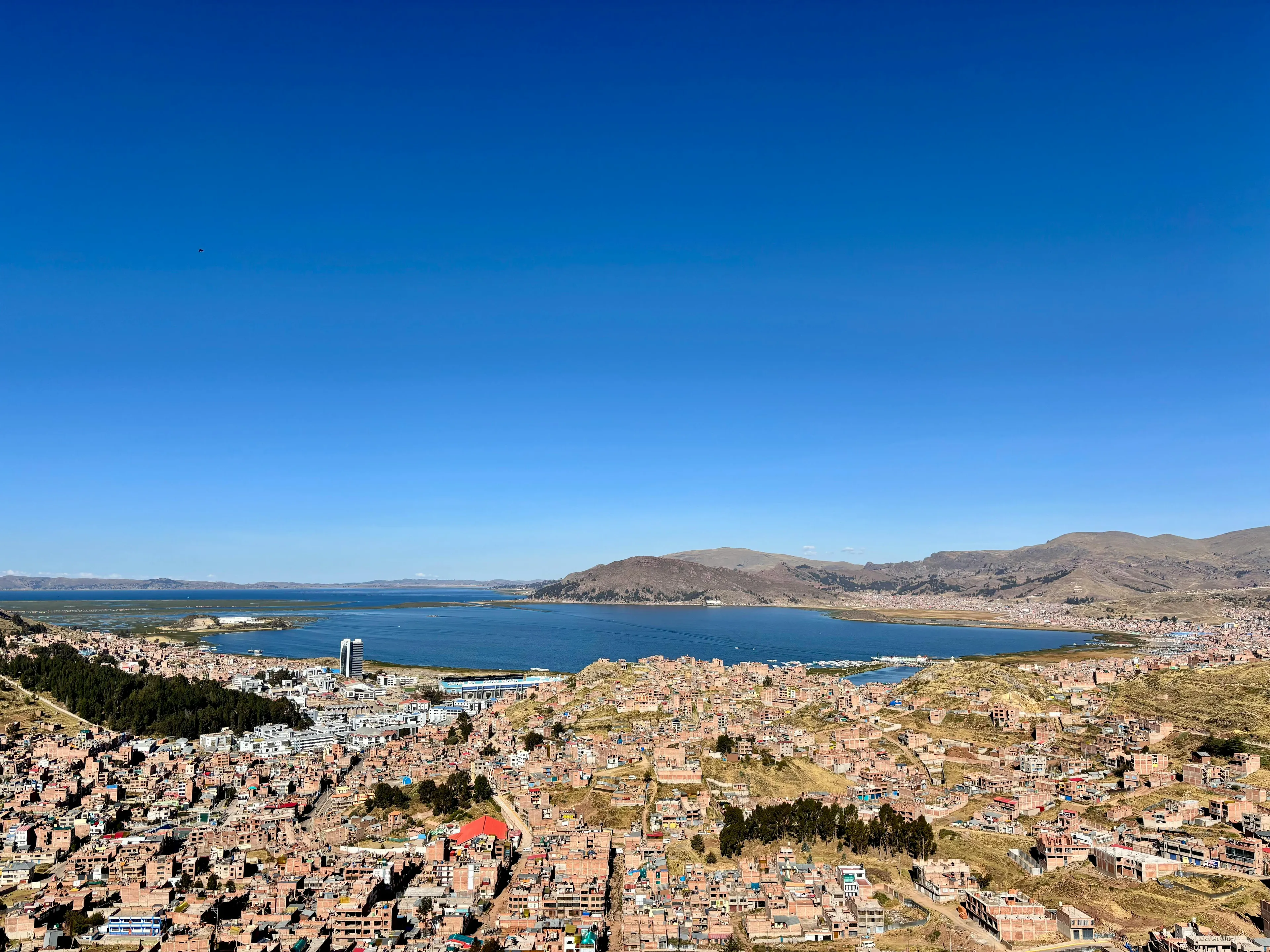 Aerial view of Lake Titicaca with surrounding mountains and urban areas under a clear blue sky.
