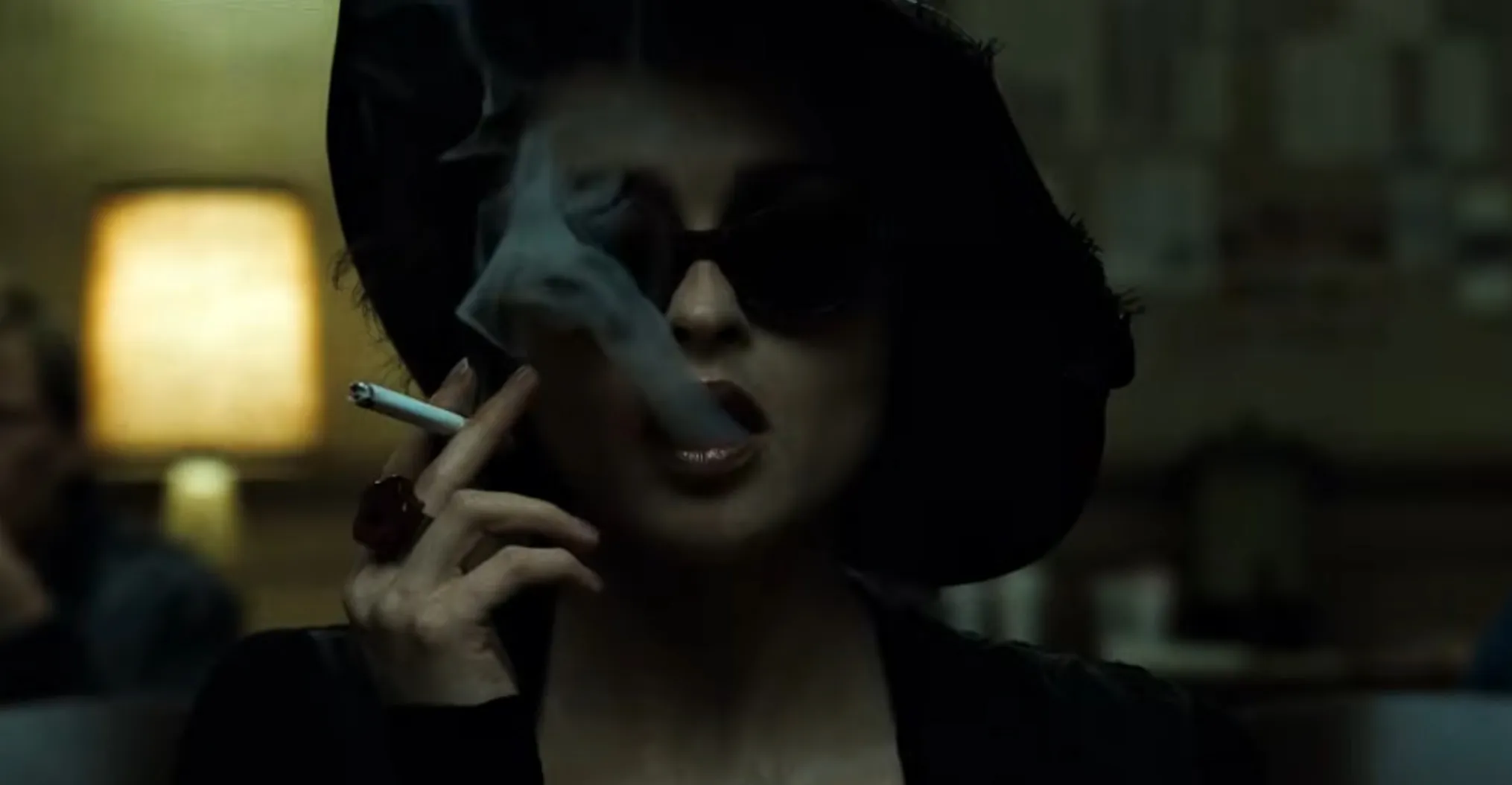 Supporting character smoking a cigarette wearing sunglasses and a black hat in a dimly lit film scene.
