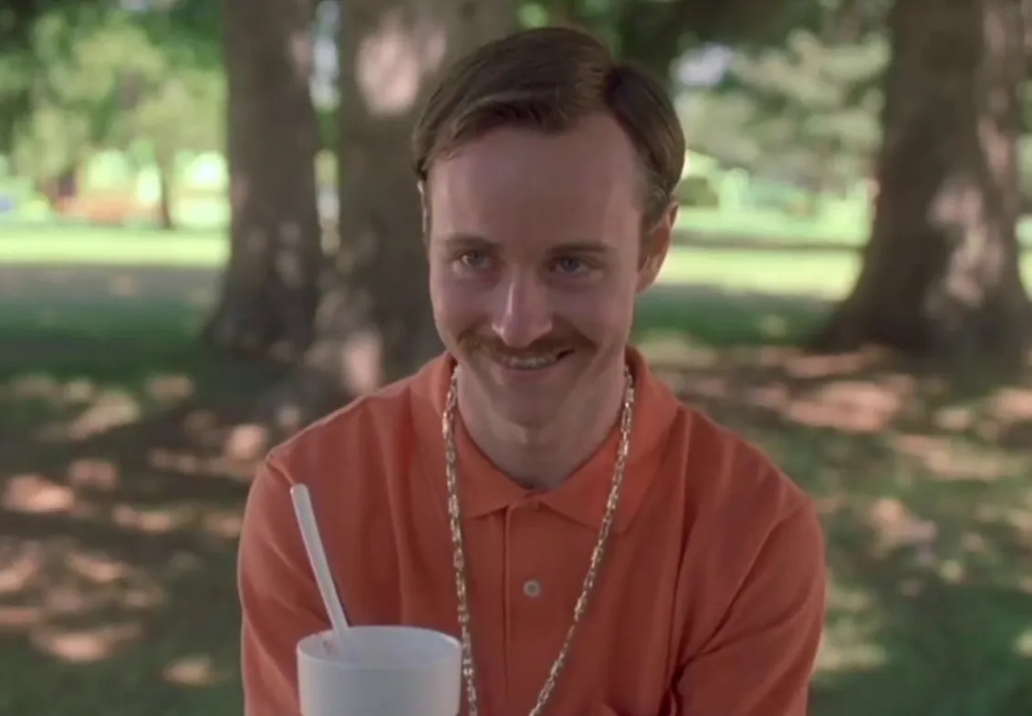 Character Rico smiling outdoors wearing an orange shirt and gold chain related to Kip in Napoleon Dynamite.