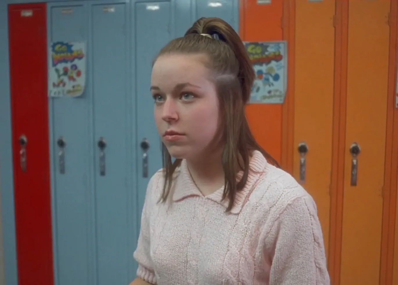 Teen girl in a light pink sweater standing in front of colorful school lockers, related to Rico uncle character in Napoleon Dynamite Deb.