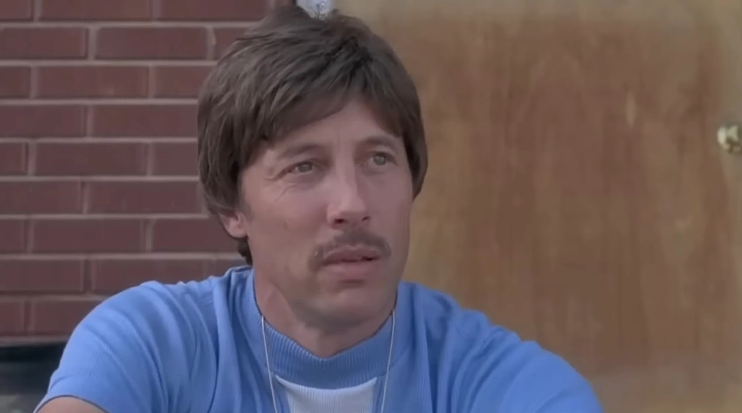 Supporting character Rico from Napoleon Dynamite with a mustache, wearing a blue shirt, sitting outdoors near a brick wall and door.