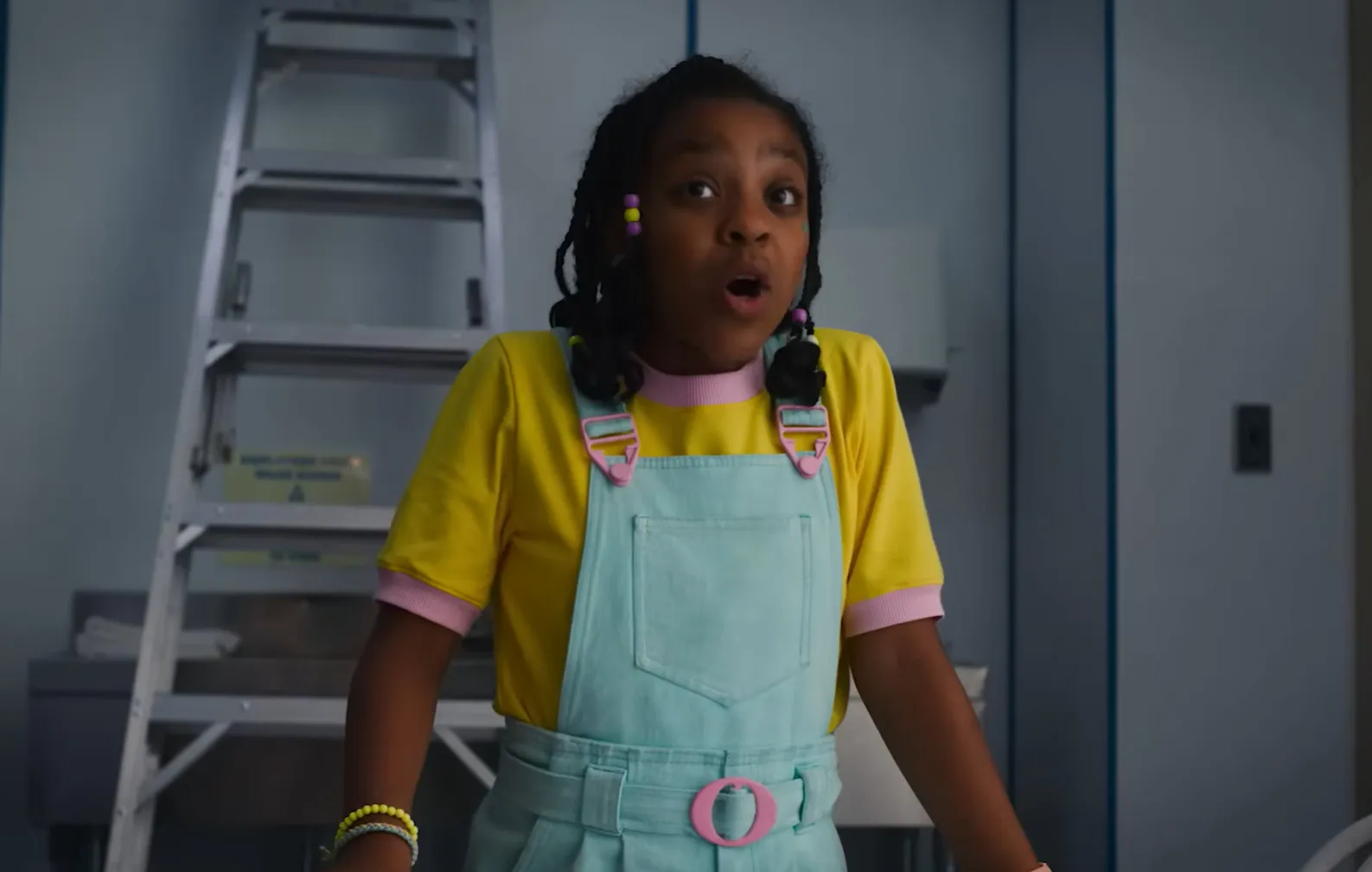 Erica Sinclair from Stranger Things wearing yellow shirt and light blue overalls with colorful beads in hair.