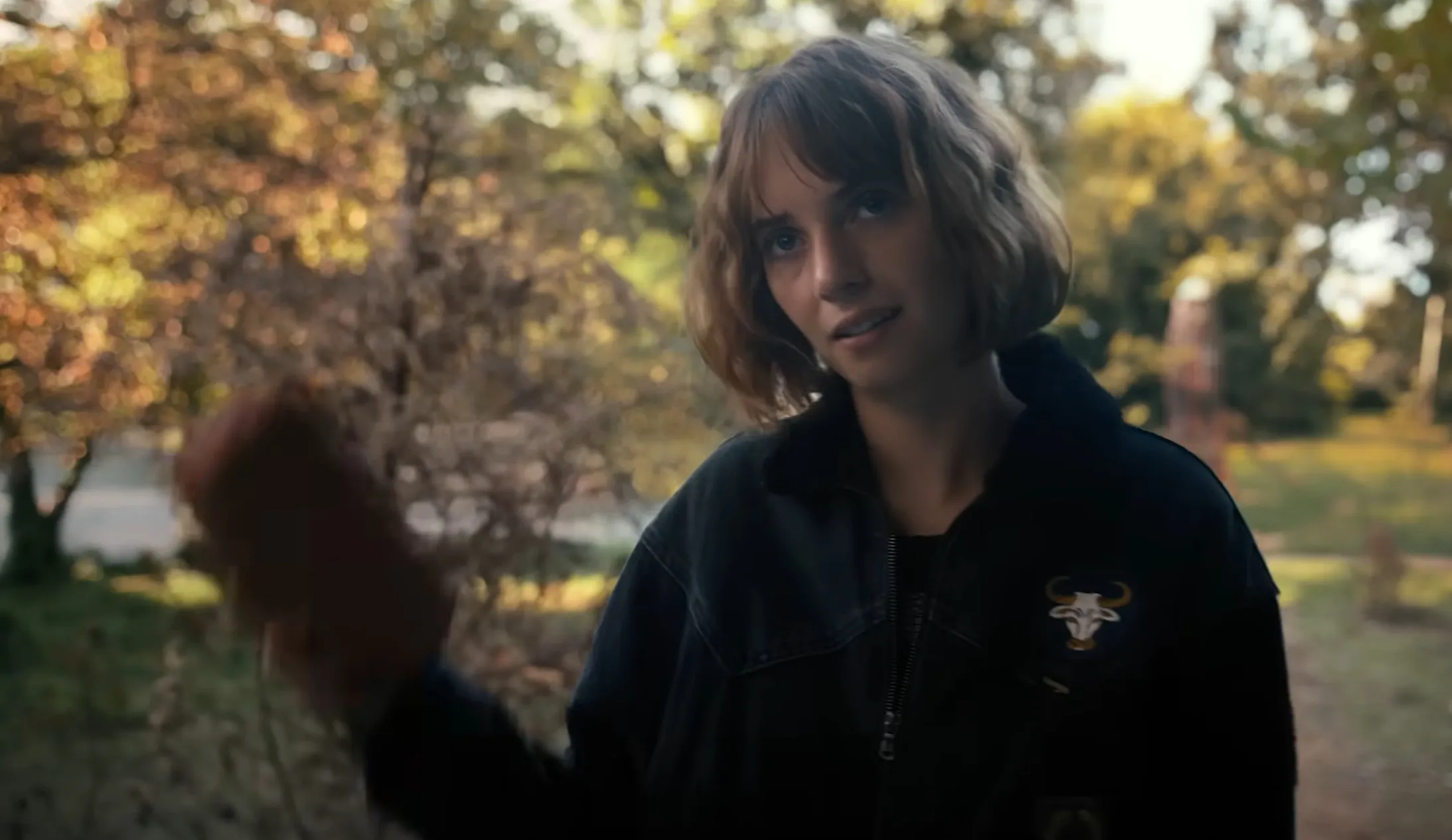 Robin Buckley from Stranger Things outdoors wearing a dark jacket with a patch, holding an object in hand during autumn.