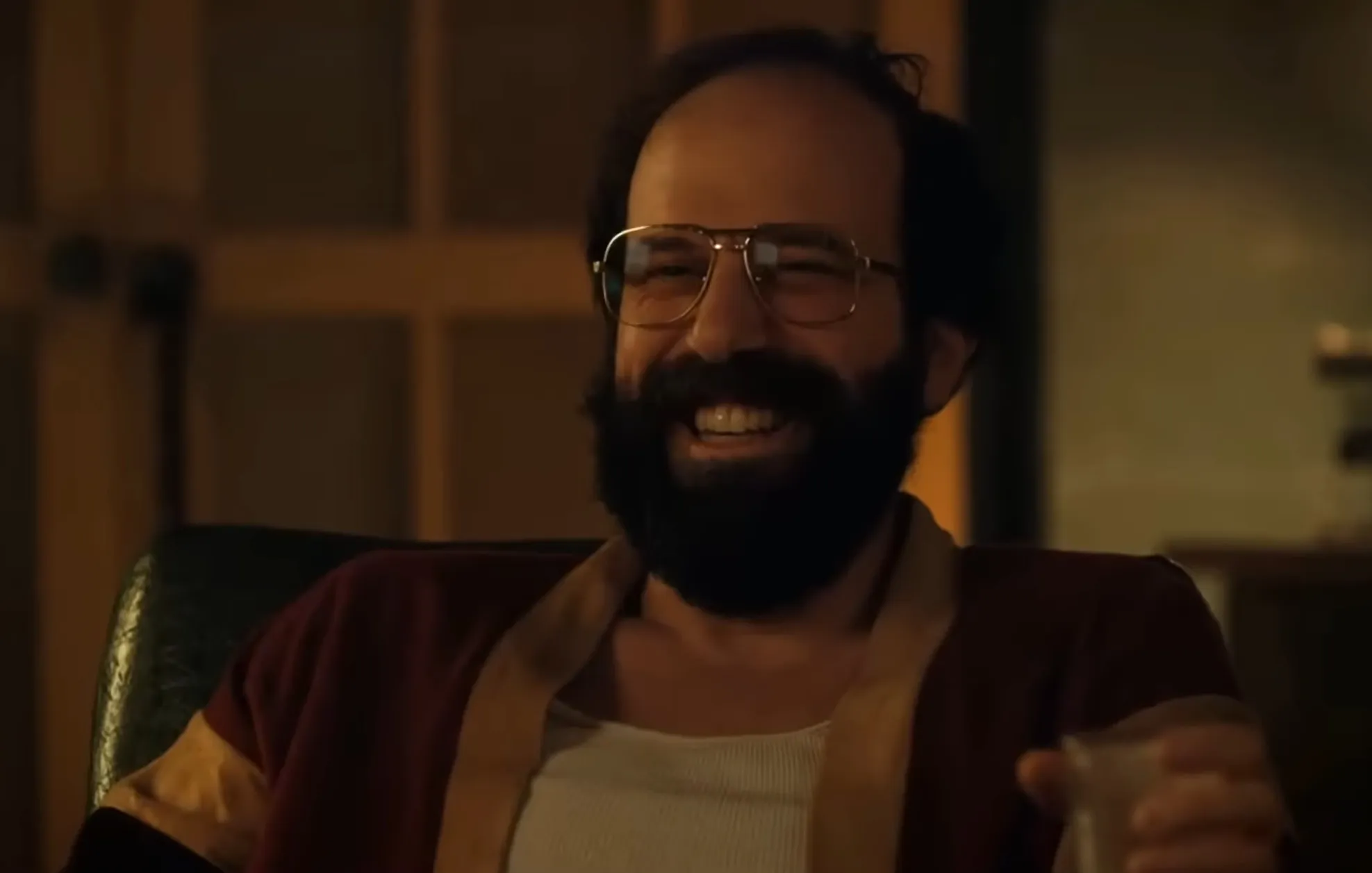Murray Bauman from Stranger Things smiling, wearing glasses and a red shirt while holding a drink in a dimly lit room