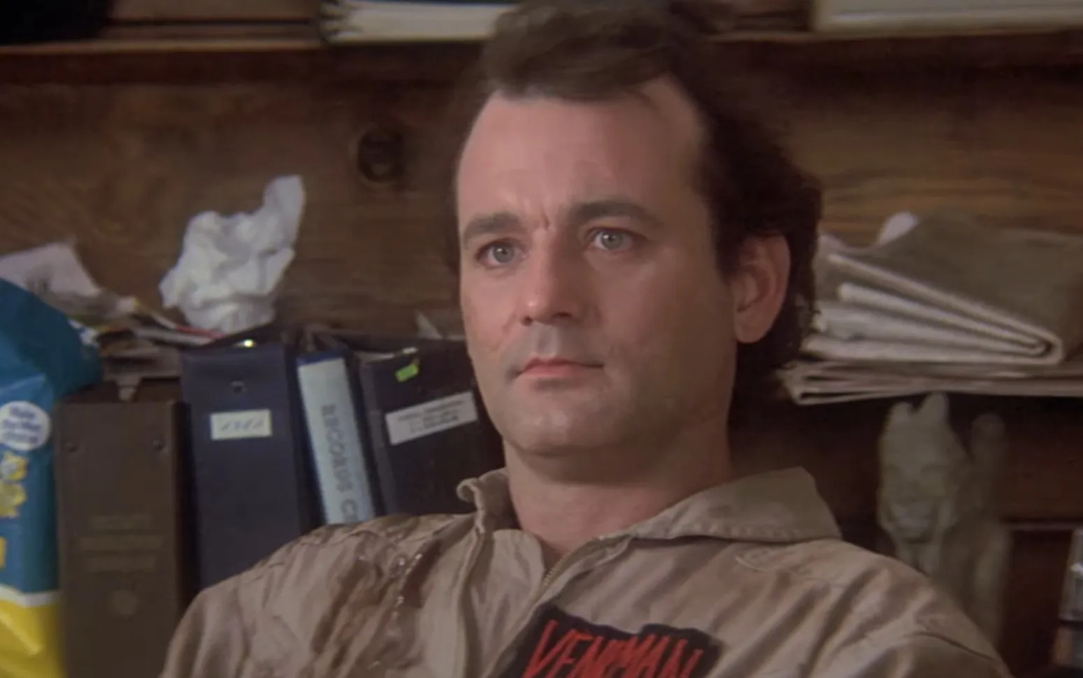 Peter Venkman in Ghostbusters uniform sitting indoors with office supplies and folders behind him.