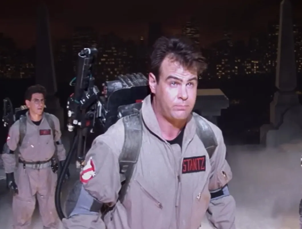 Two Ghostbusters in gray uniforms with proton packs at night in a city setting, related to character love interest theme.