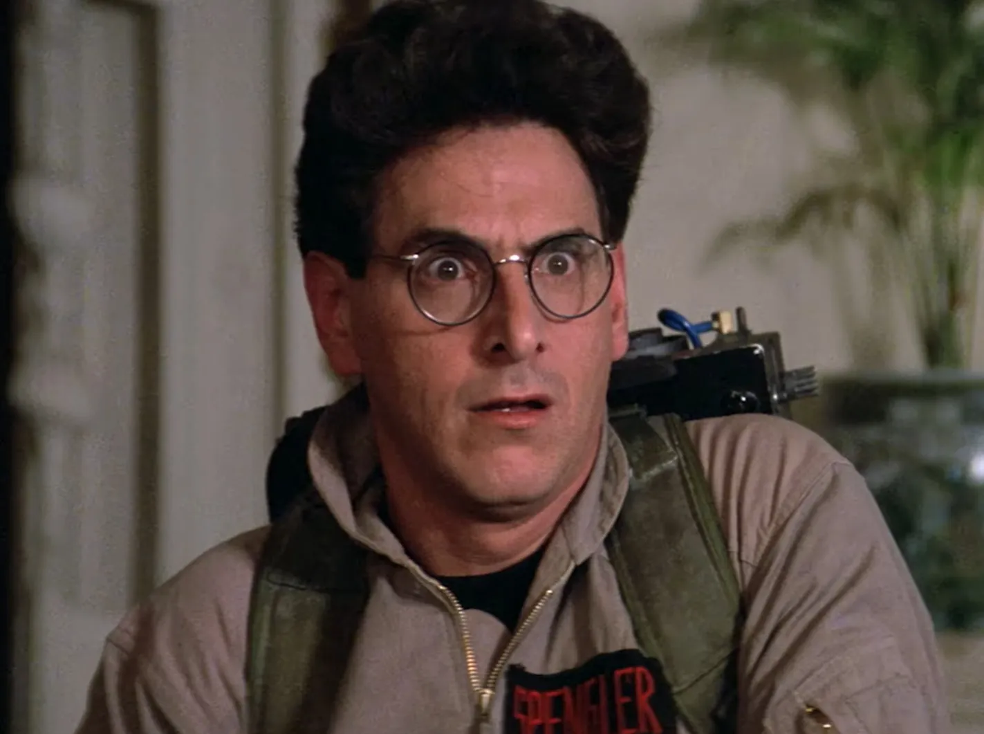 Egon Spengler in Ghostbusters uniform with round glasses and surprised expression indoors with blurred background.