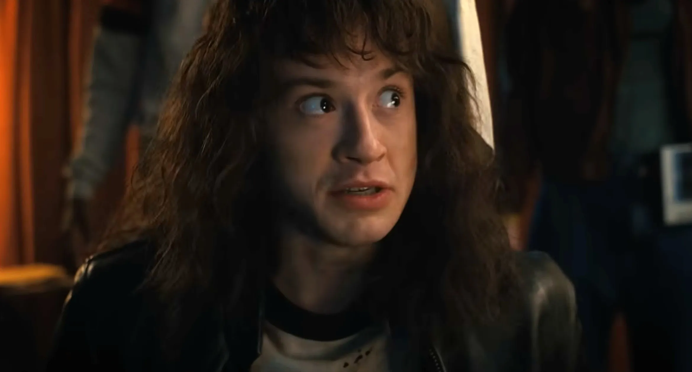 Eddie Munson from Stranger Things with long curly hair wearing a leather jacket in a dimly lit room.