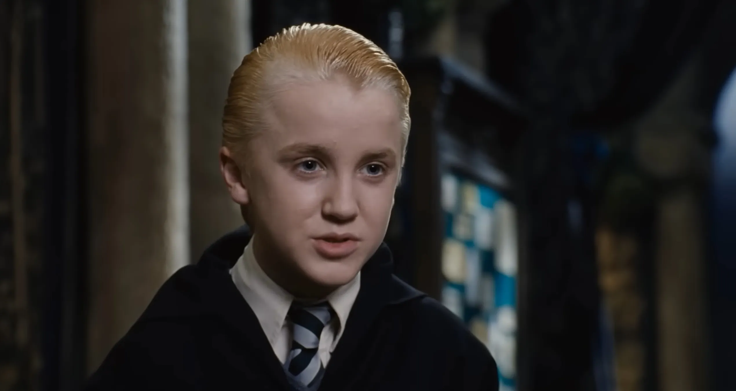 Blond supporting character from the Harry Potter films wearing Hogwarts uniform and striped tie indoors.
