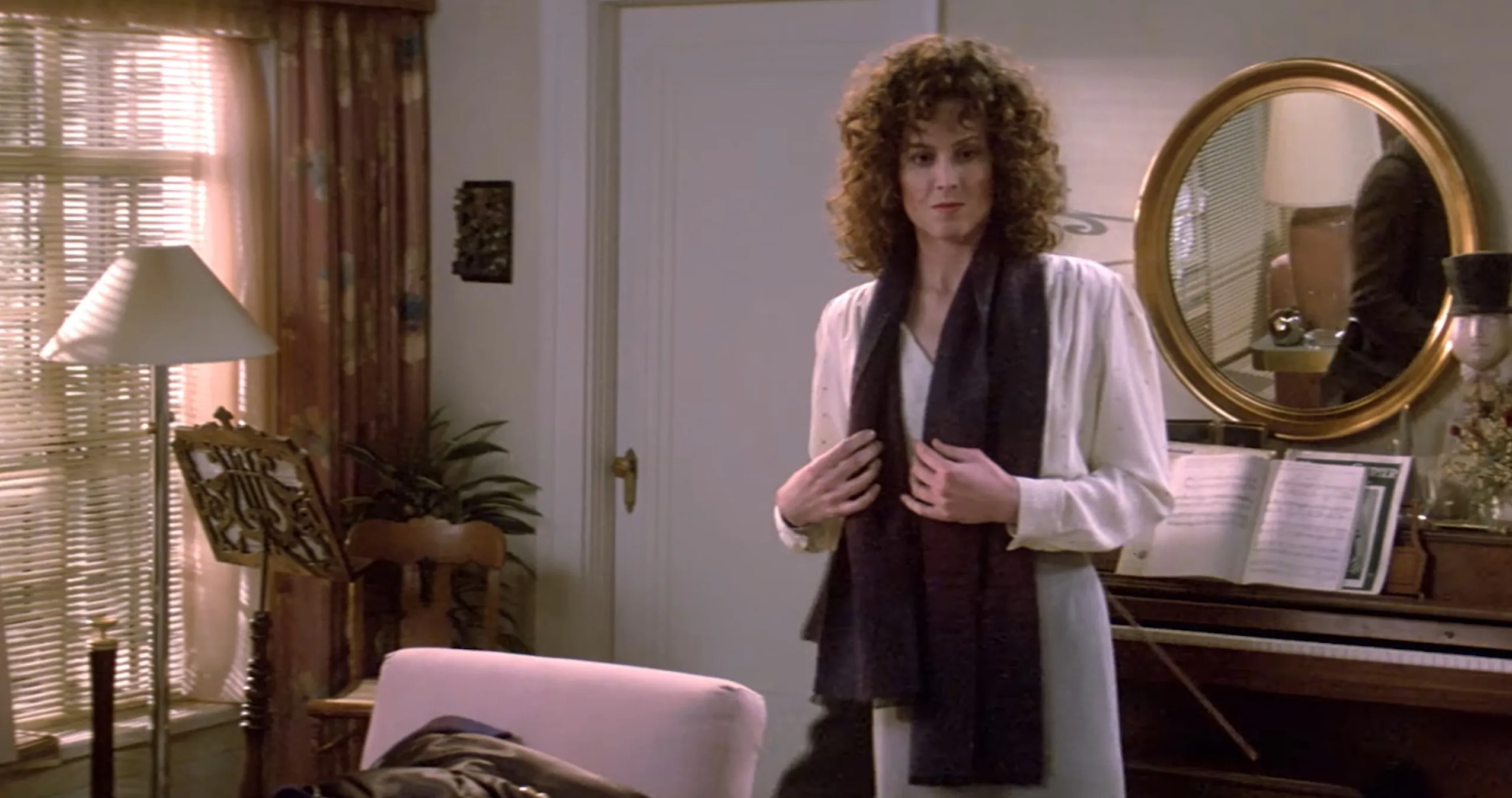 Supporting character with curly hair standing in a living room near a piano with sheet music and a round mirror behind.