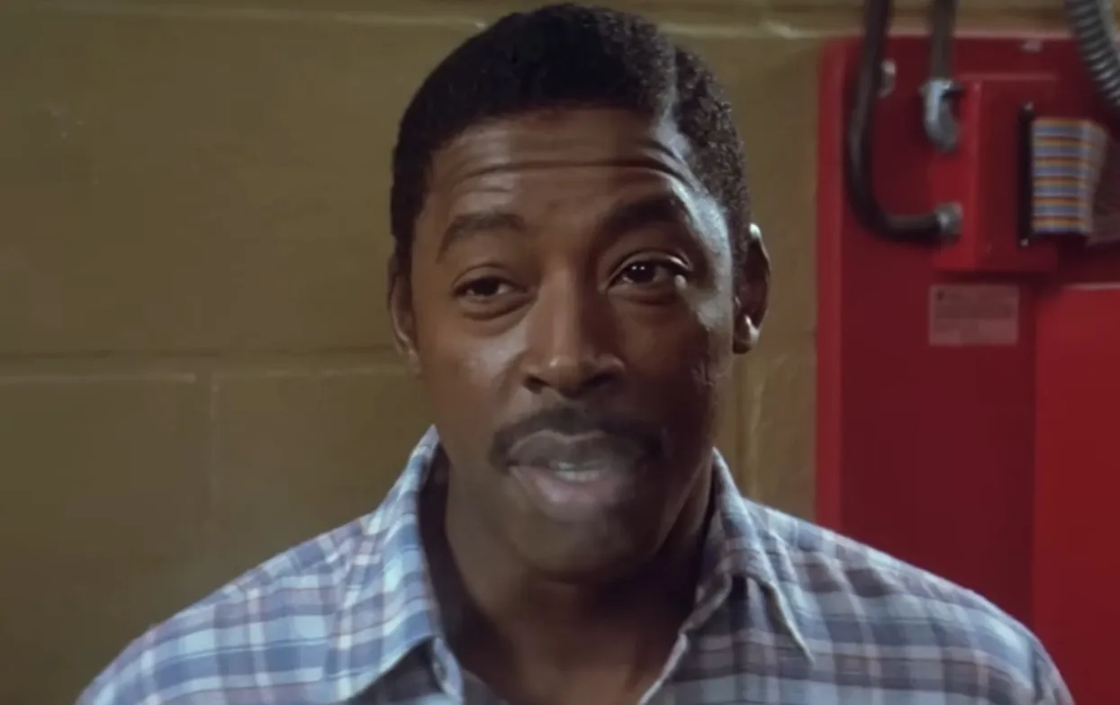 Winston Zeddemore character close-up wearing a plaid shirt in an indoor setting with a red panel in the background