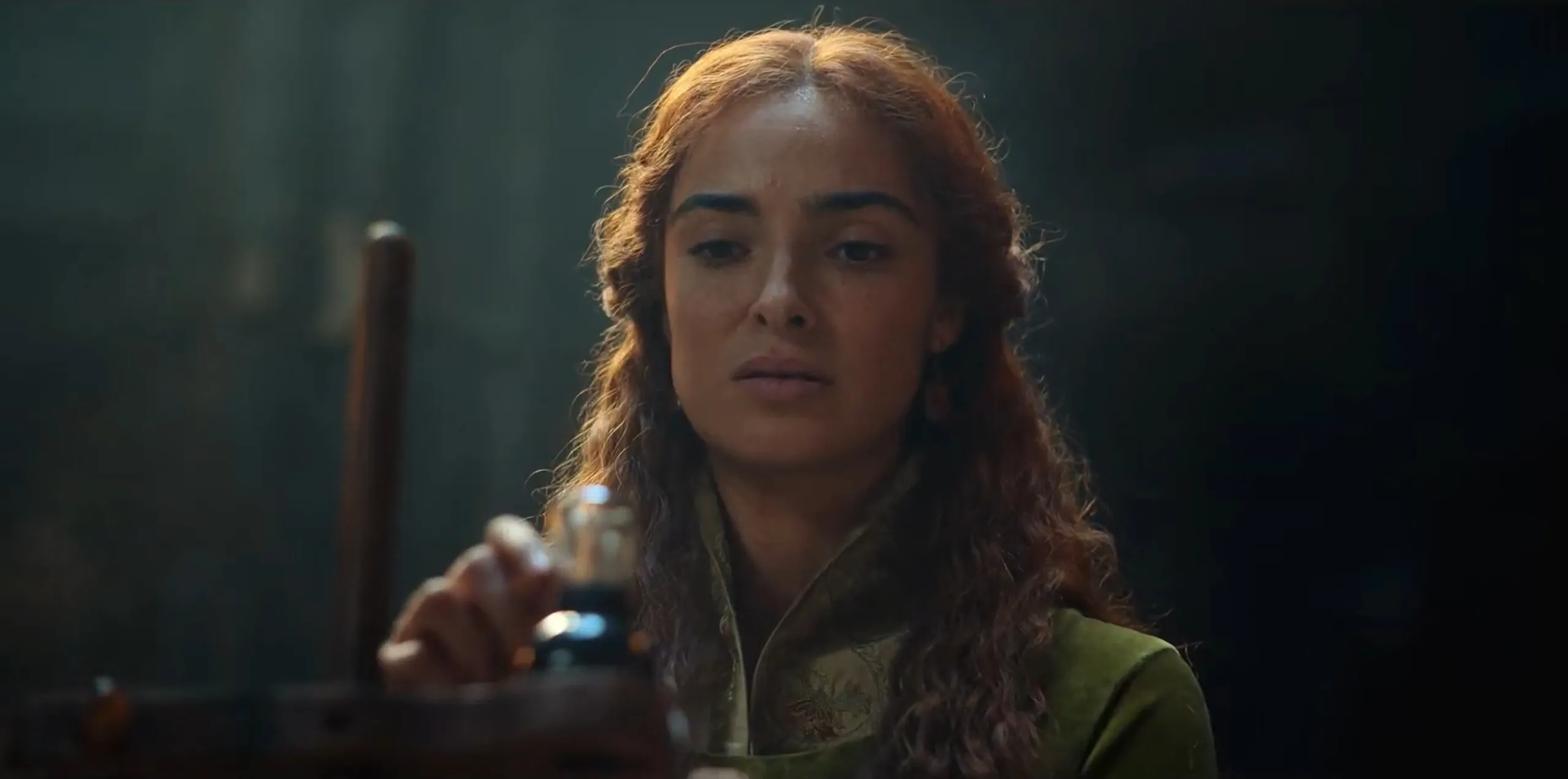 Supporting character from The Witcher holding a bottle, acting as mentor to a main cast character in a dark setting.