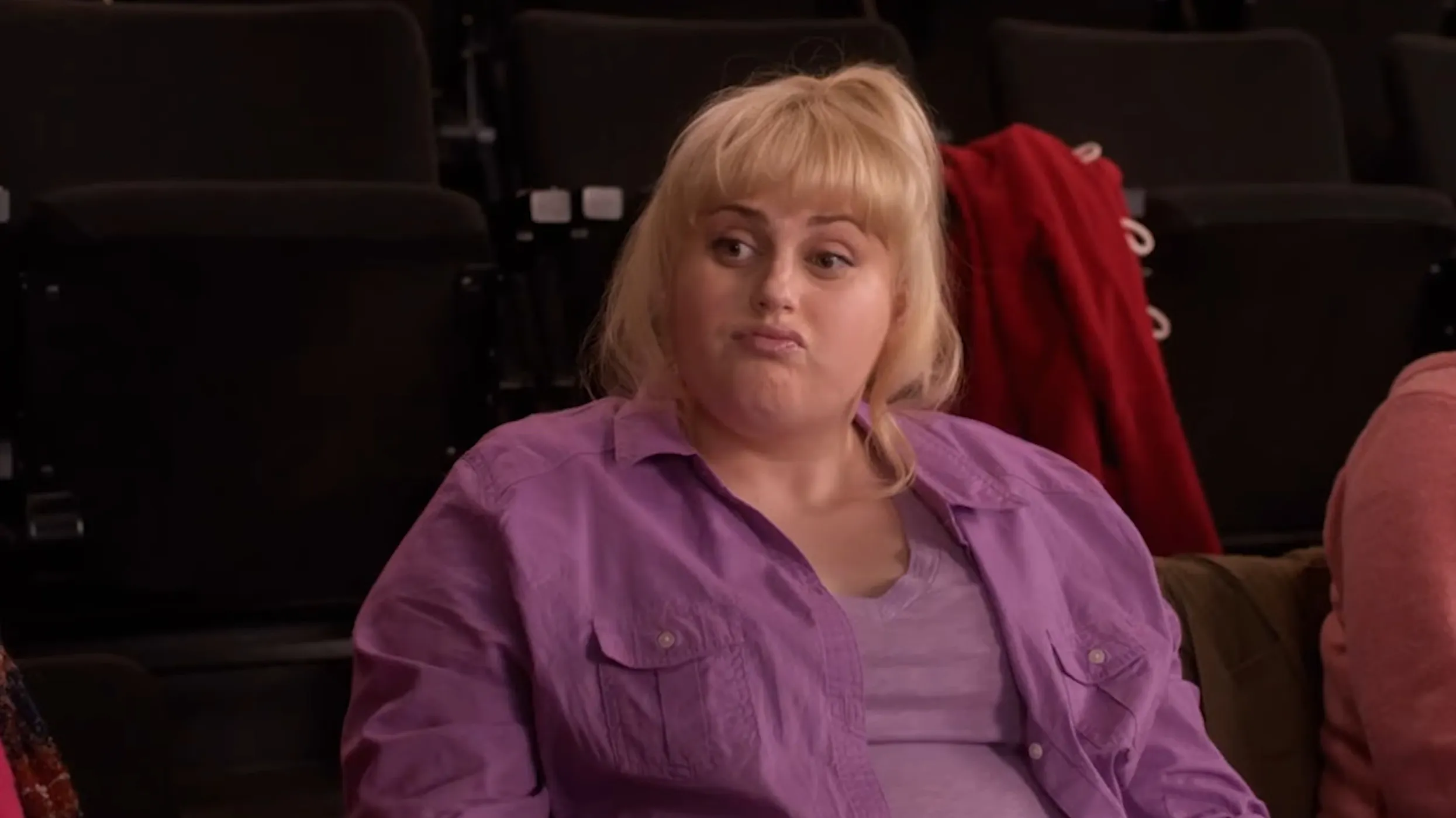 Supporting character from a musical wearing a purple shirt sitting in a theater with a skeptical facial expression.