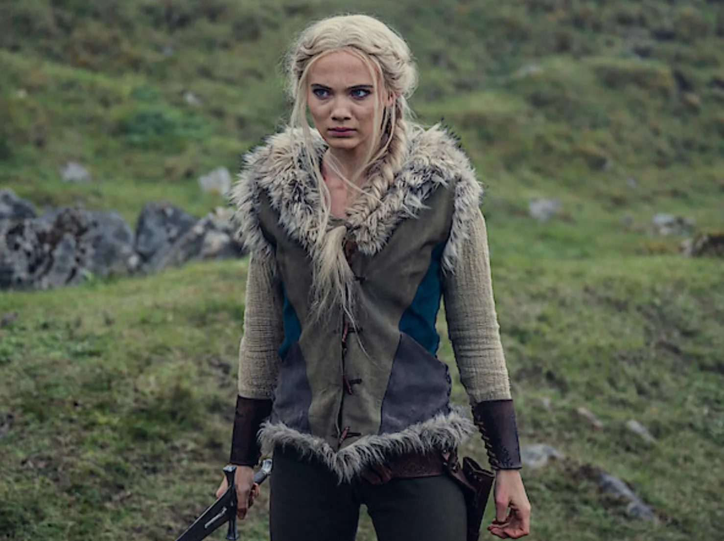 The Witcher character Ciri's mentor in a fur-lined medieval outfit holding a small weapon in an outdoor setting