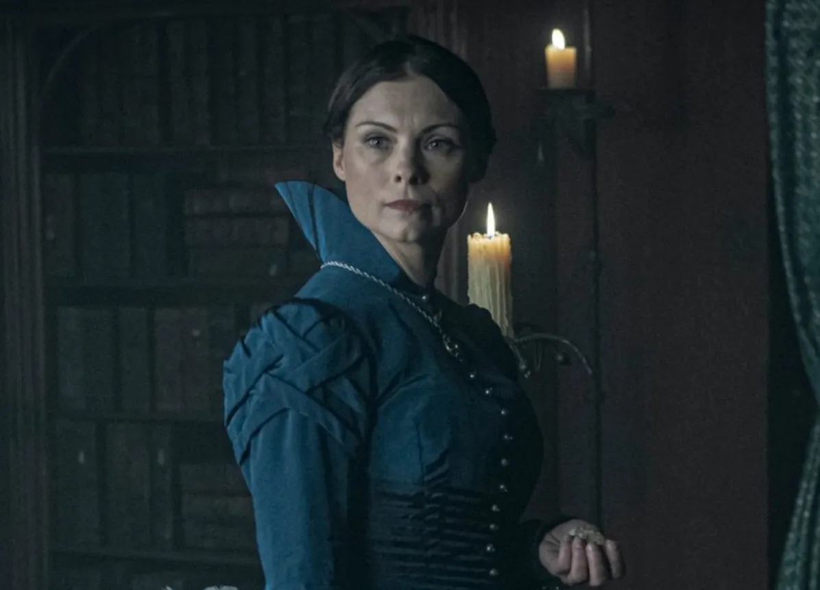 Tissaia from The Witcher wearing a dark blue dress, standing in a dimly lit room with candles behind her.