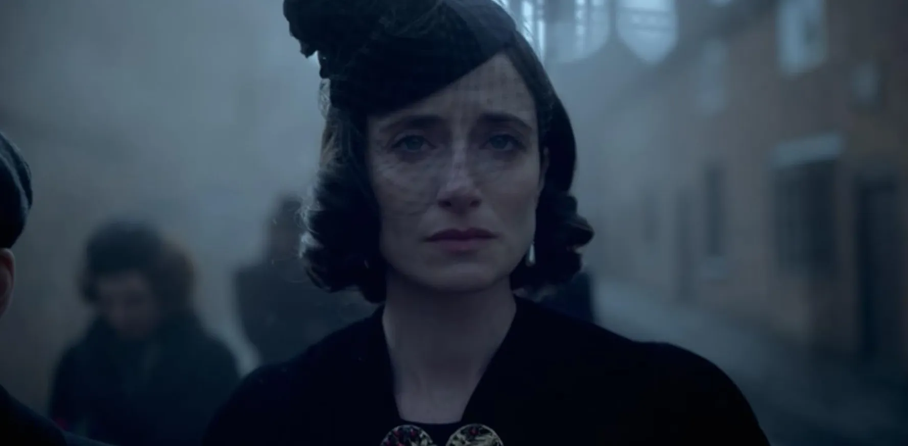 Woman in vintage attire with a netted hat, looking somber, scene hinting at supporting character in a show.