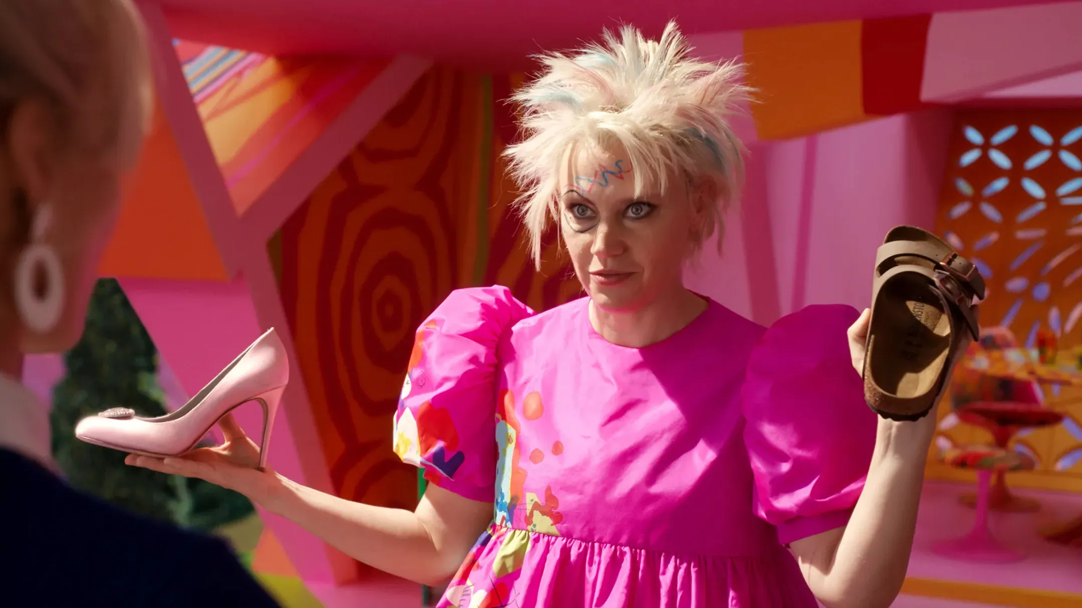 Quirky supporting character in bright pink dress holding two different shoes in a colorful film scene.